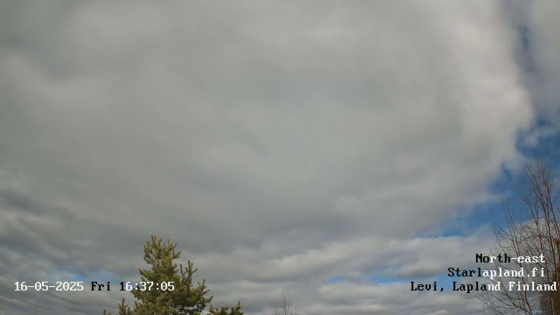 A mostly cloudy sky with some blue sky visible, and a few small trees are visible in the bottom corners of the frame.