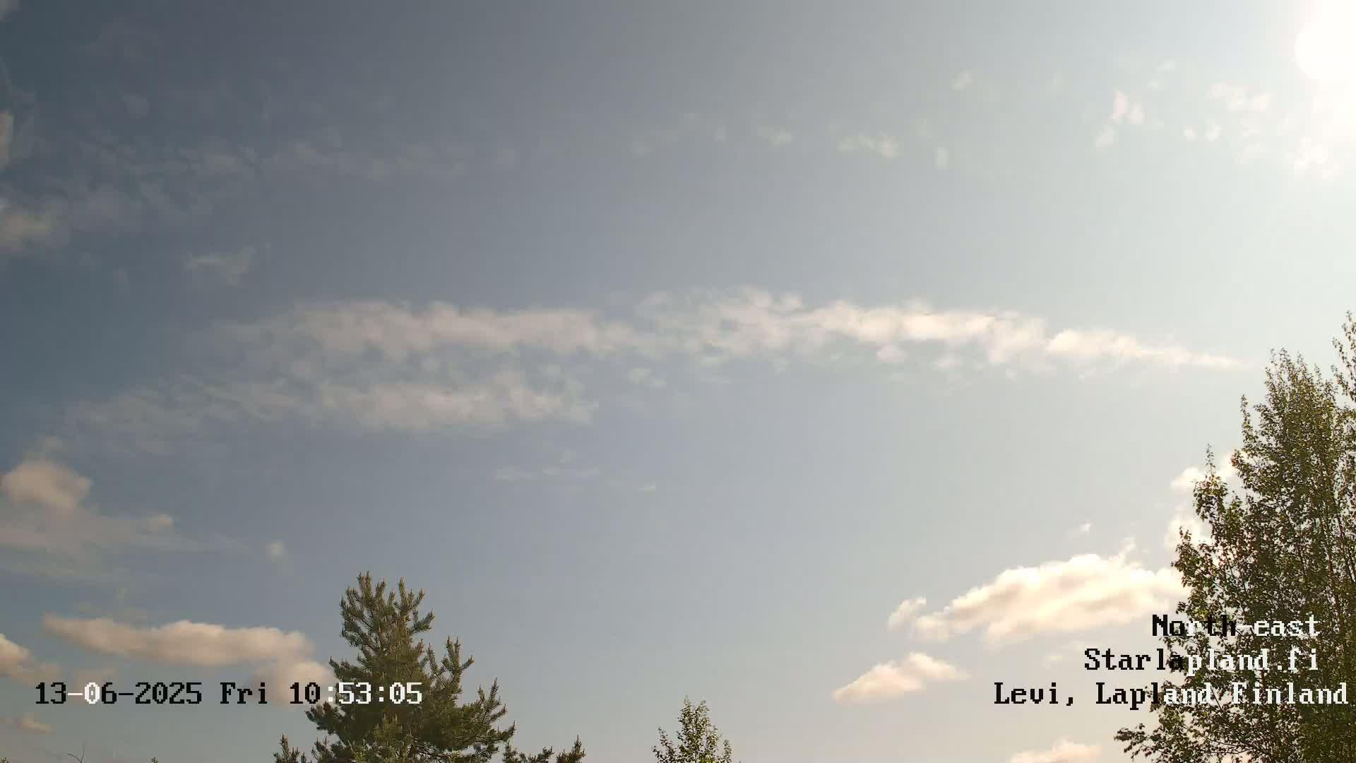 A partly cloudy sky with scattered cumulus clouds is visible above a landscape featuring several coniferous and deciduous trees.