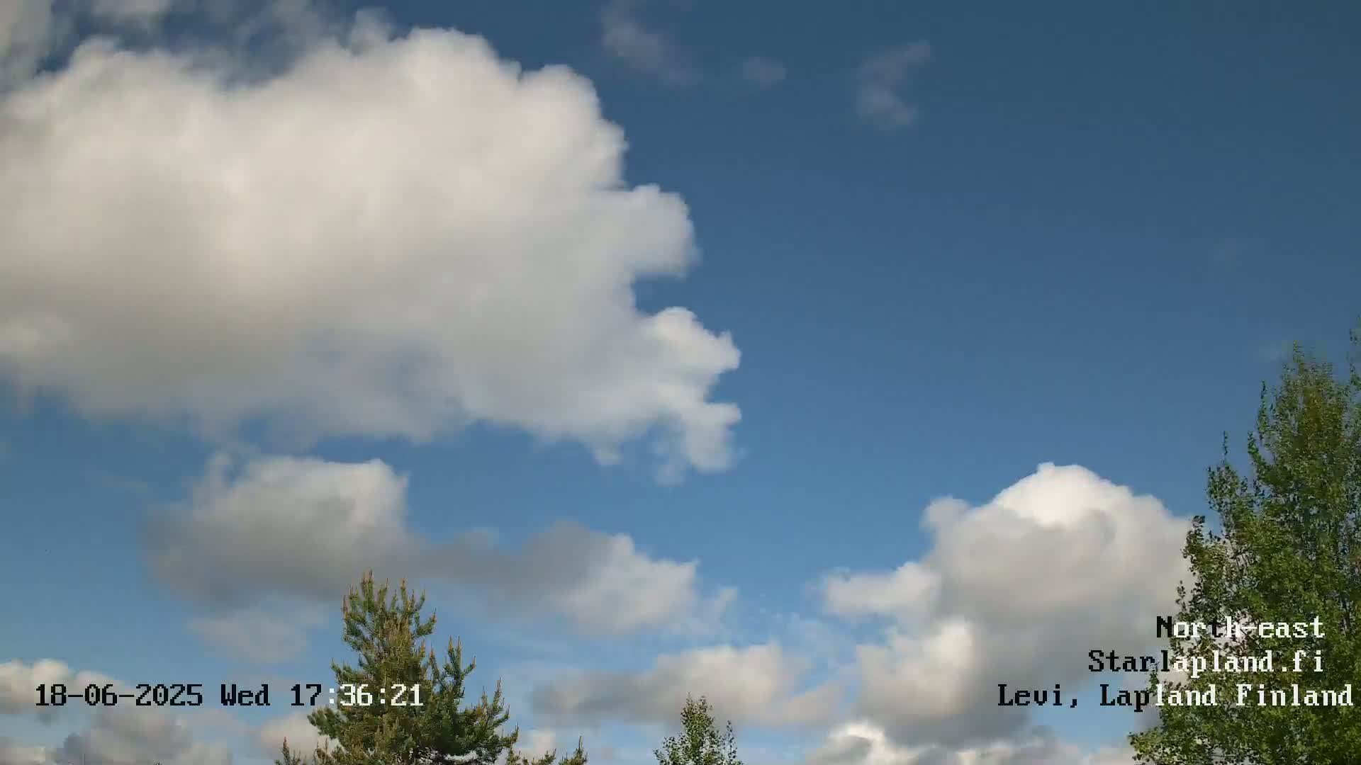 A partly cloudy sky with fluffy white cumulus clouds against a clear blue background, and some green trees are visible at the bottom.