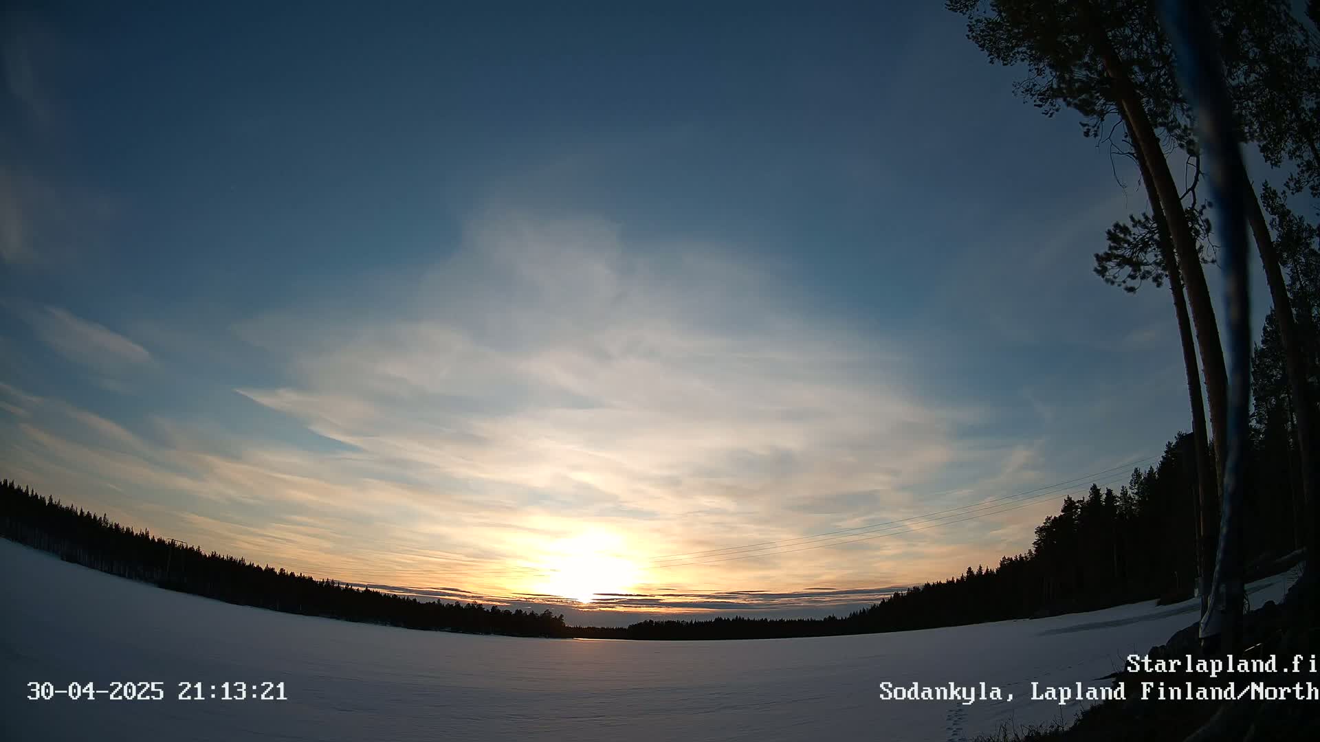 A snowy, flat landscape is illuminated by a low setting sun under a mostly clear, light-blue sky with thin, wispy clouds.