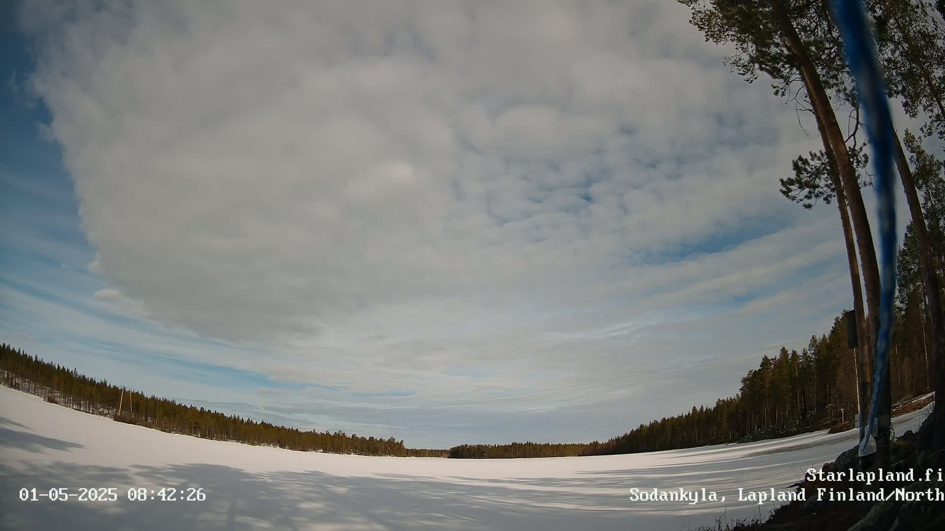 A mostly cloudy sky over a snow-covered, frozen lake bordered by a dark pine forest.