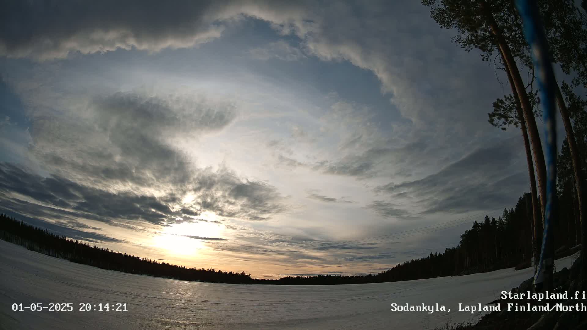 A wide, low-angle view shows a mostly cloudy sunset over a snow-covered landscape with a dark forest in the distance.
