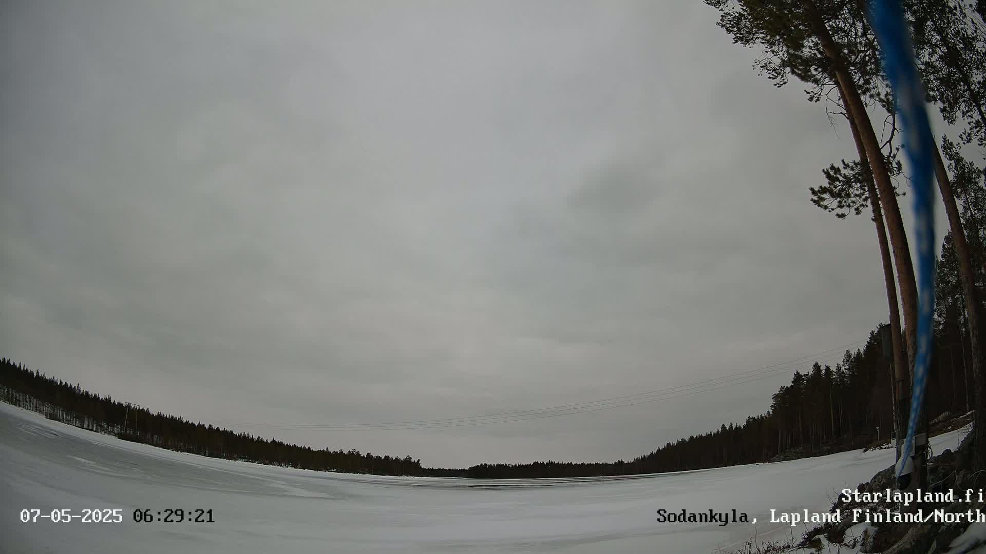 A wide shot shows a snow-covered landscape under a cloudy, overcast sky, with a dark forest bordering a frozen body of water.