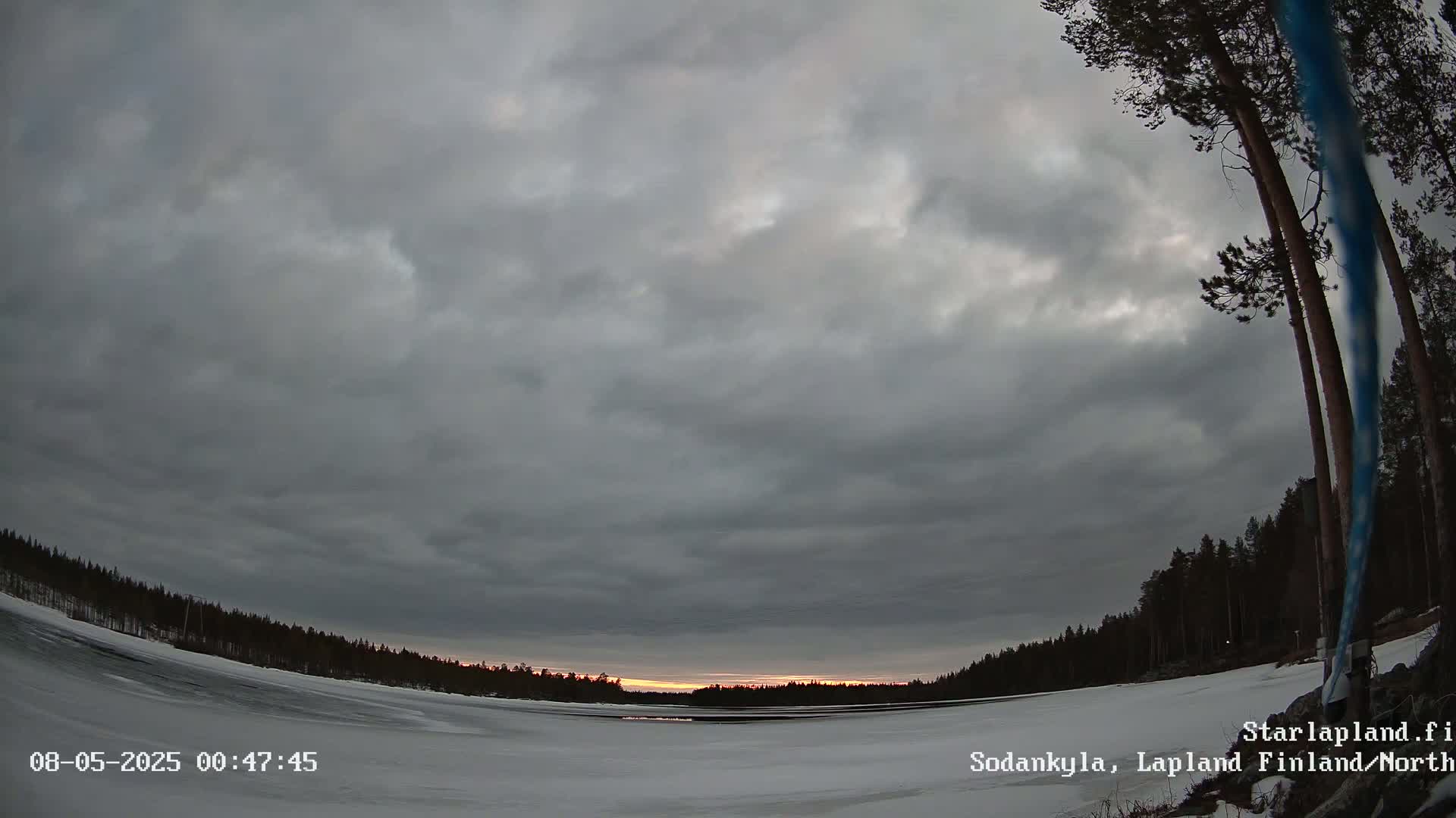 A mostly cloudy, twilight sky over a snow-covered frozen lake and dark evergreen forest.