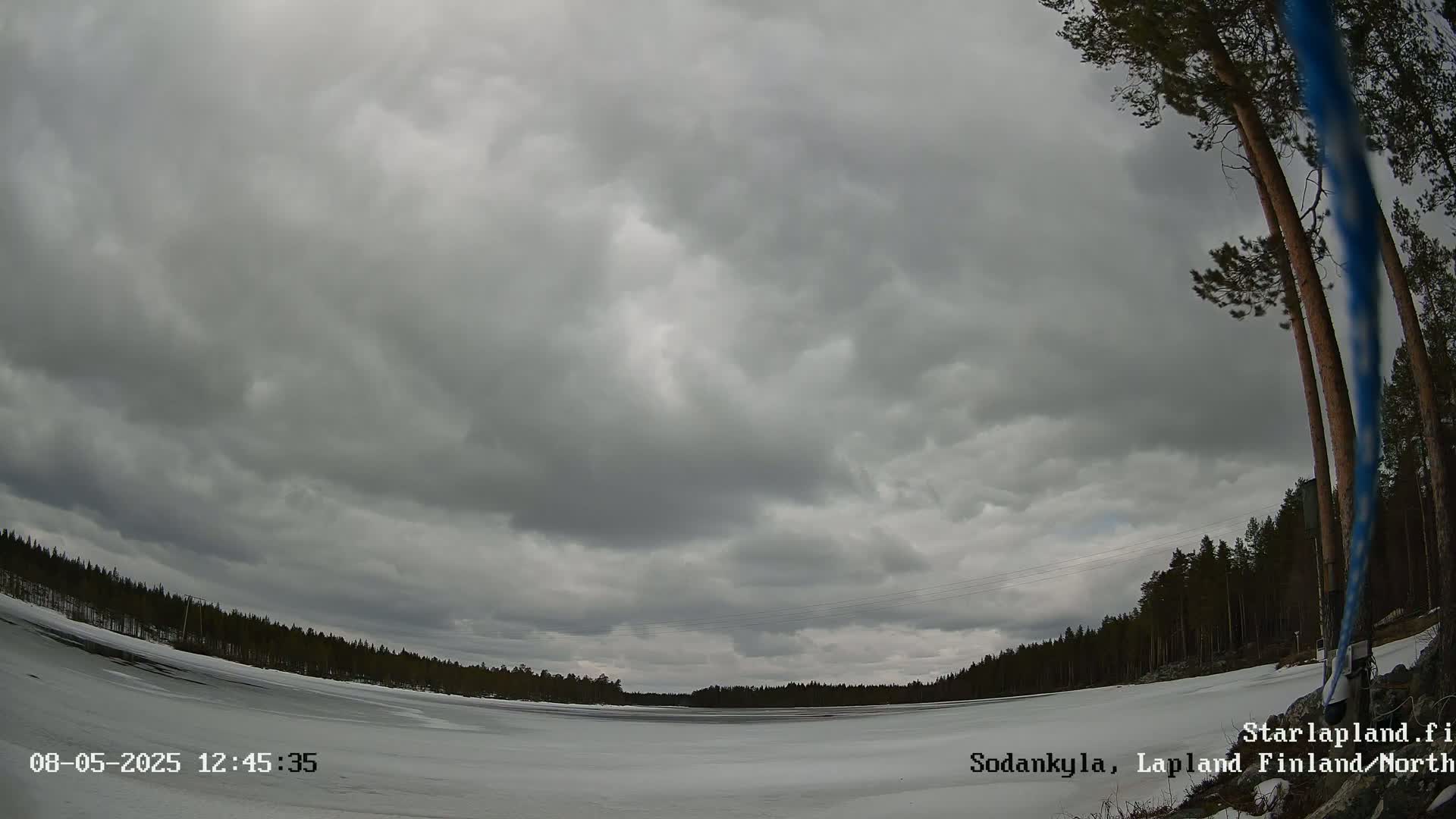 A mostly cloudy sky over a snow-covered frozen lake, bordered by dark evergreen trees.