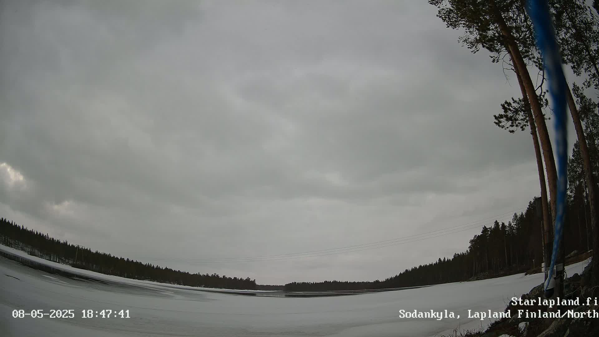 A mostly cloudy, overcast sky covers a snow-covered landscape with a partially frozen lake and dark evergreen trees in the background.