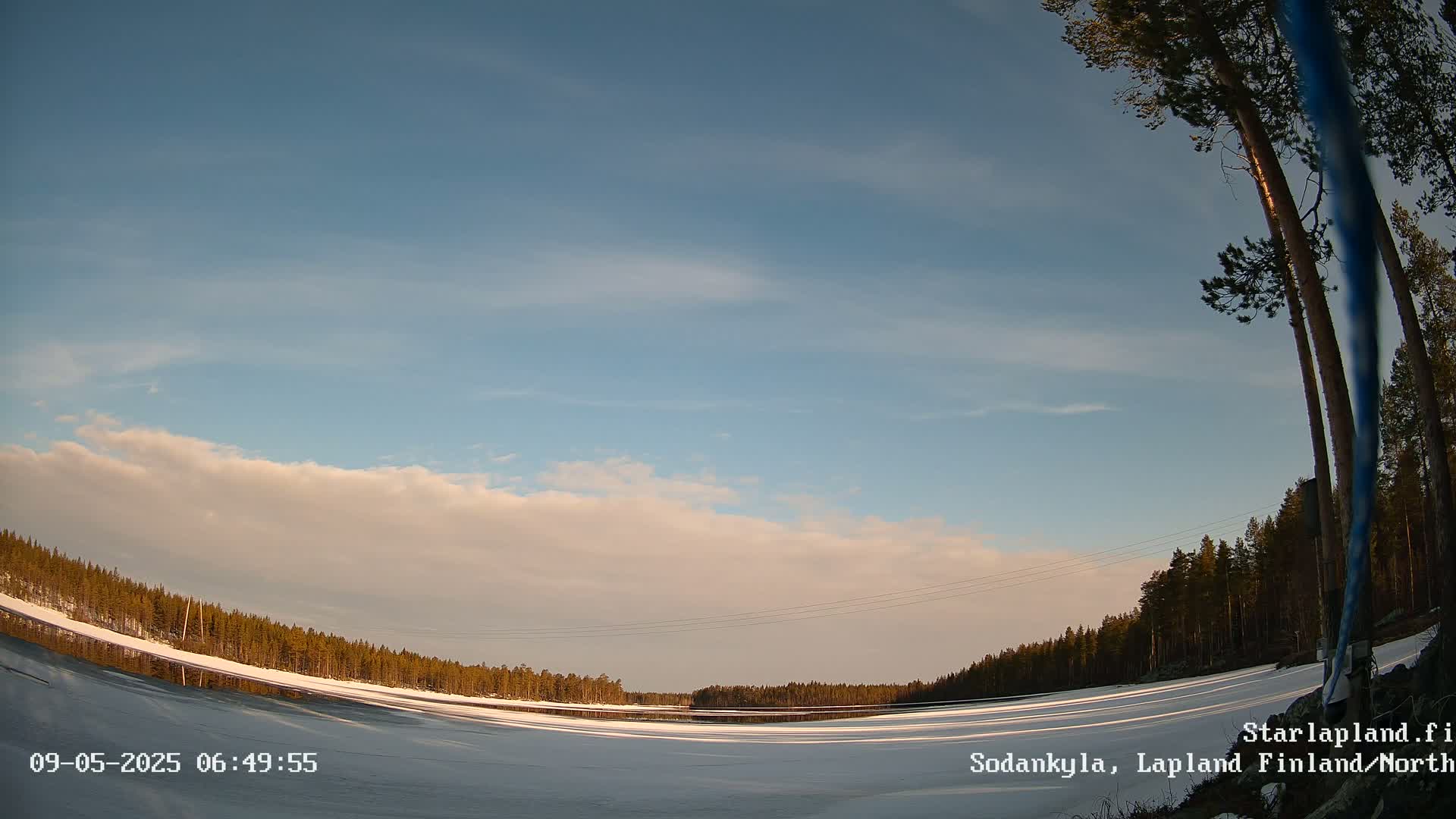 A partly cloudy, bright sky overlooks a snow-covered frozen lake and a dark evergreen forest.
