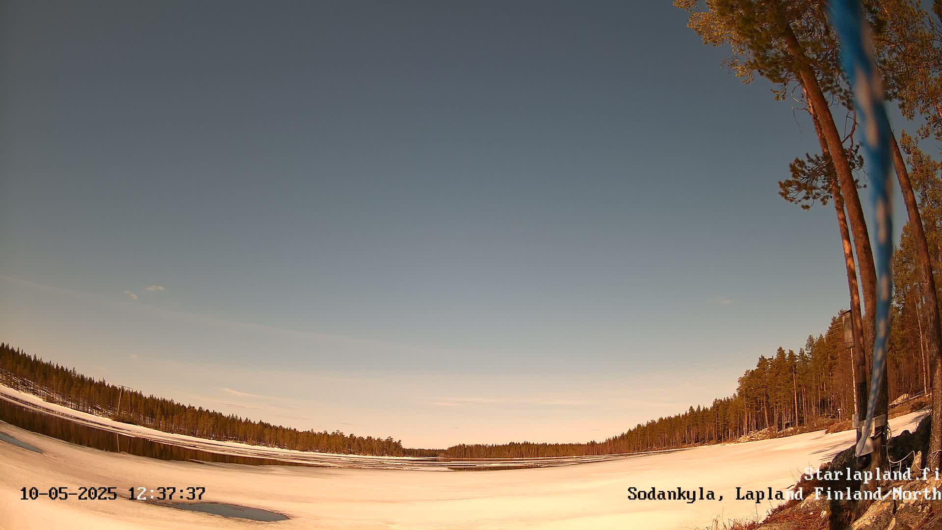 A mostly clear sky overlooks a partially frozen lake surrounded by a snow-covered shoreline and coniferous forest.