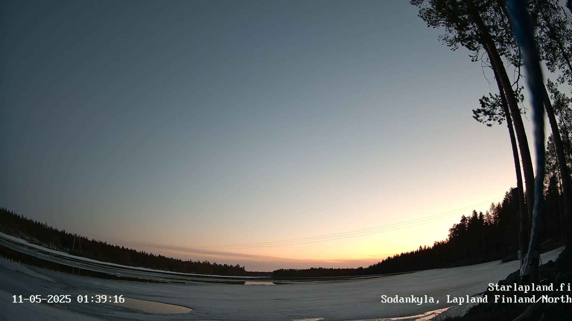 A twilight sky over a snow-covered landscape with a partially frozen body of water and silhouetted trees in the foreground and background.