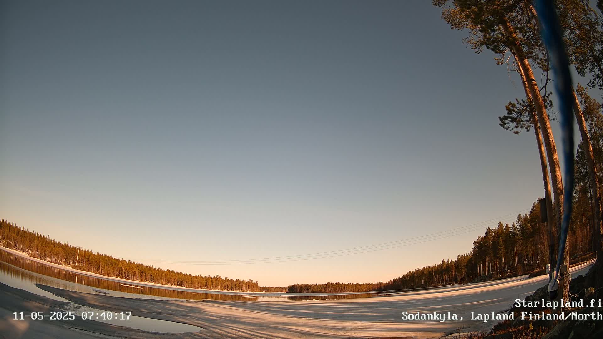 A partly frozen lake is surrounded by a snow-covered shoreline and coniferous trees under a clear, twilight sky.