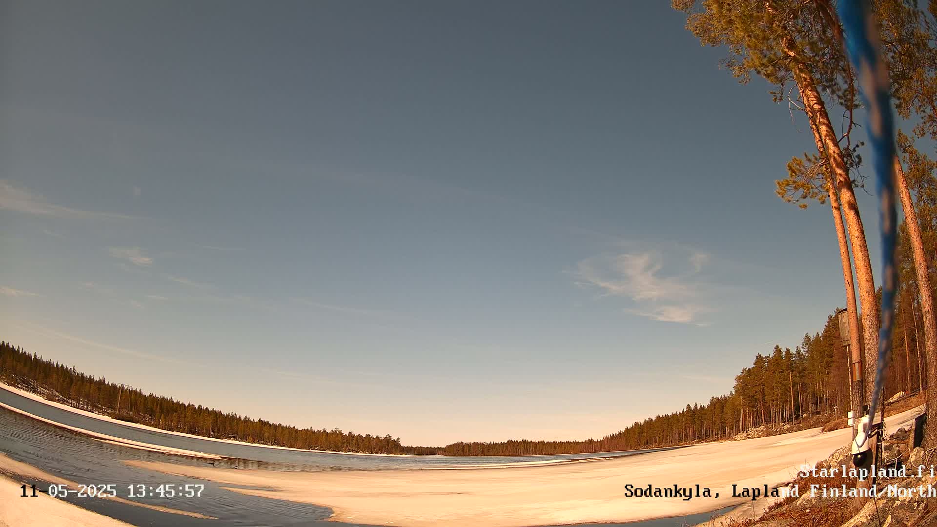 A partly frozen lake is surrounded by a snow-covered shoreline and a dark pine forest under a mostly clear, light blue sky.