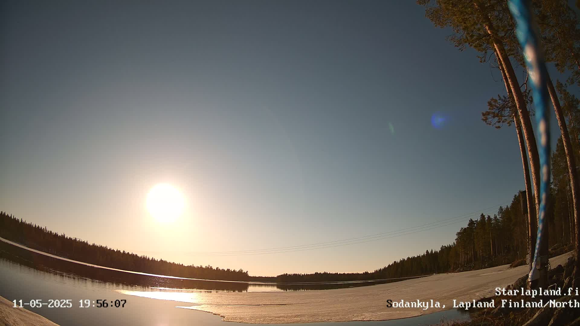 A low-angle, wide shot shows a partially frozen lake under a bright, clear sunset sky, surrounded by a dark pine forest.