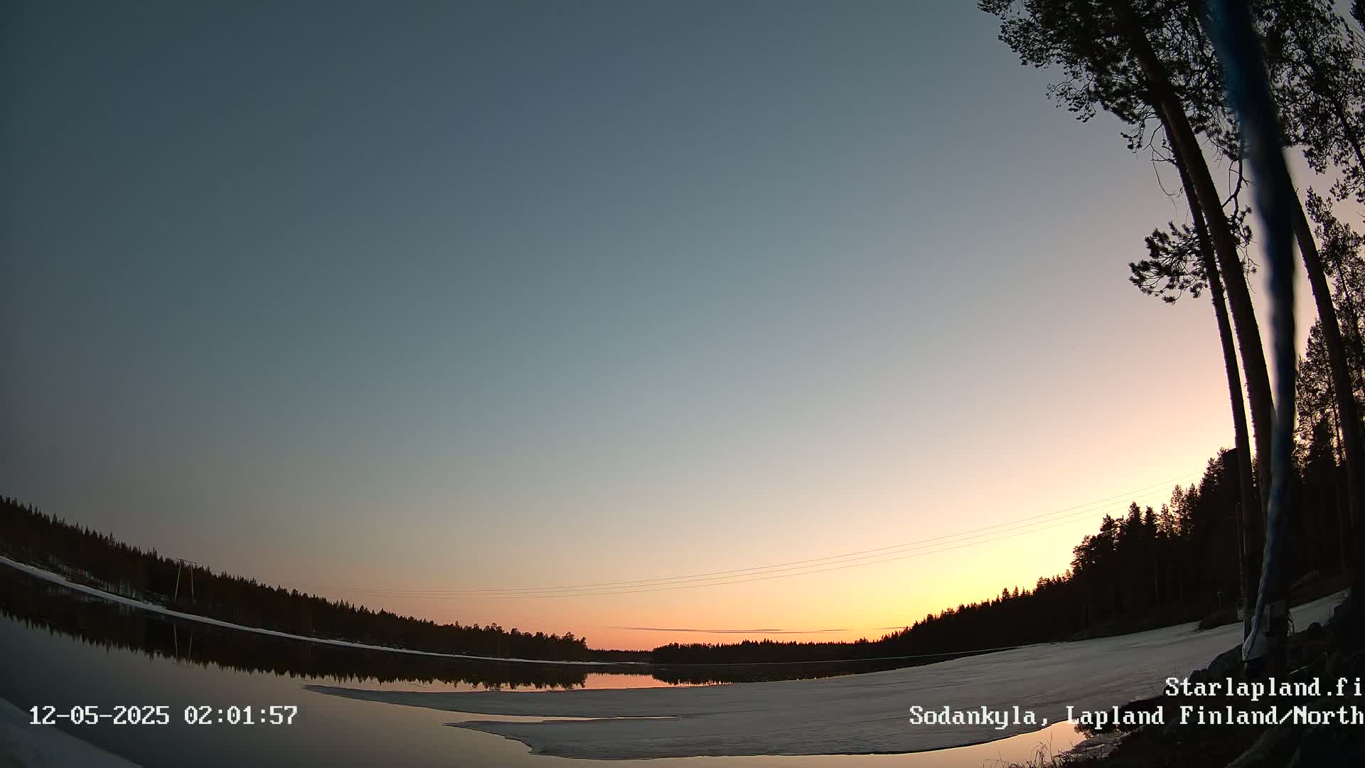 A calm, partially frozen lake reflects a sunset sky under a clear, twilight sky, bordered by dark trees and snow.
