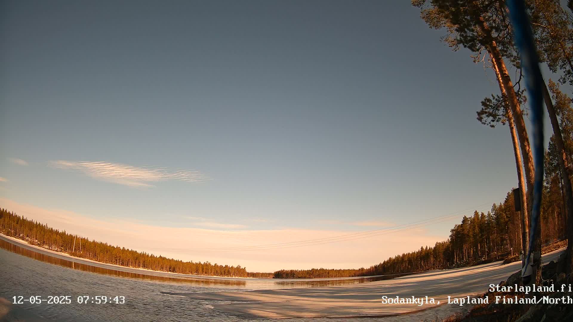 A partly frozen lake is surrounded by a snow-covered forest under a mostly clear, pale-blue sky at sunrise or sunset.