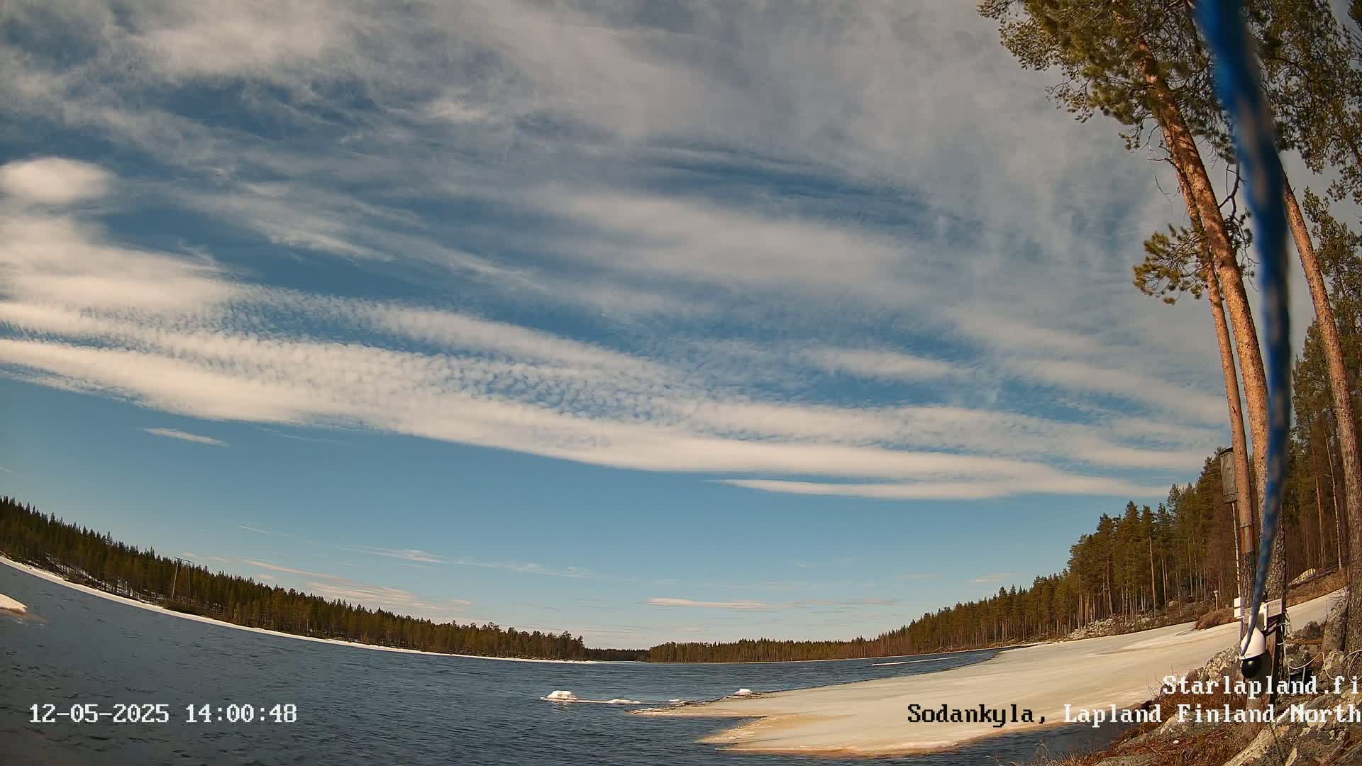 A partly frozen lake is surrounded by snow-covered banks and a coniferous forest under a mostly clear sky with thin, wispy clouds.