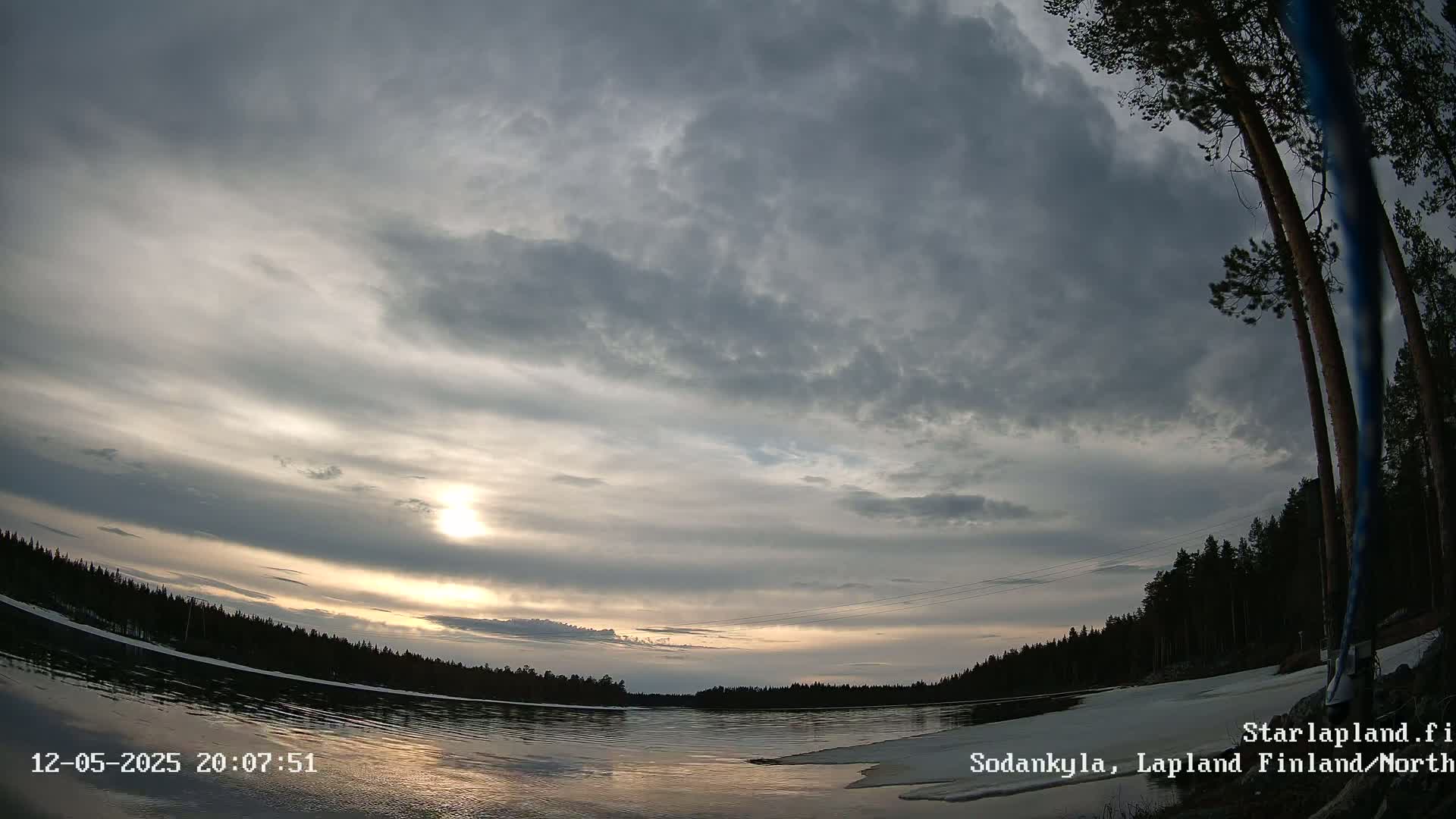 A partly cloudy sunset over a calm lake with some remaining ice along the shore, viewed from a low angle.