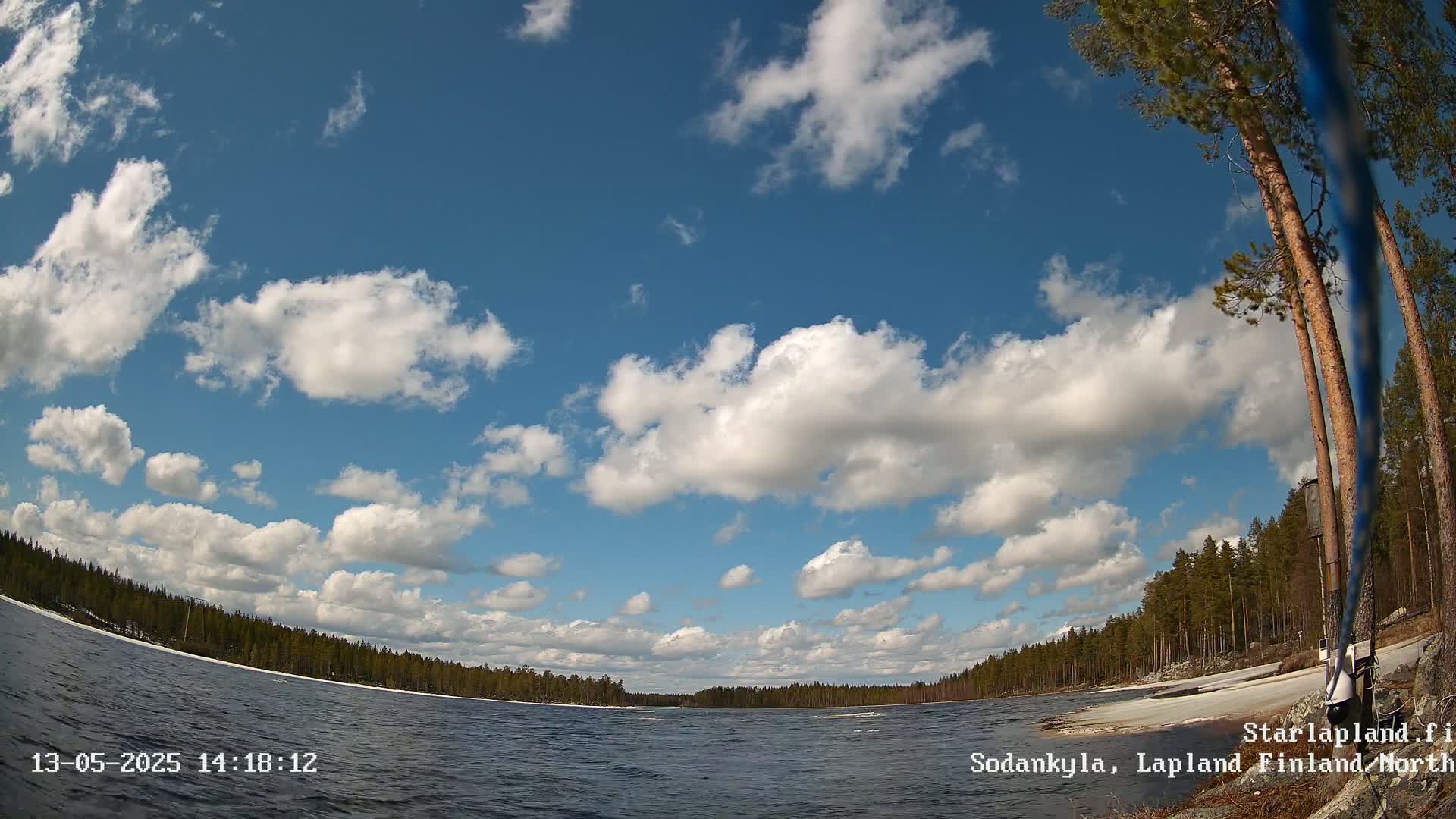 A calm lake, partially frozen near the shore, is surrounded by a coniferous forest under a mostly sunny sky with fluffy white clouds.