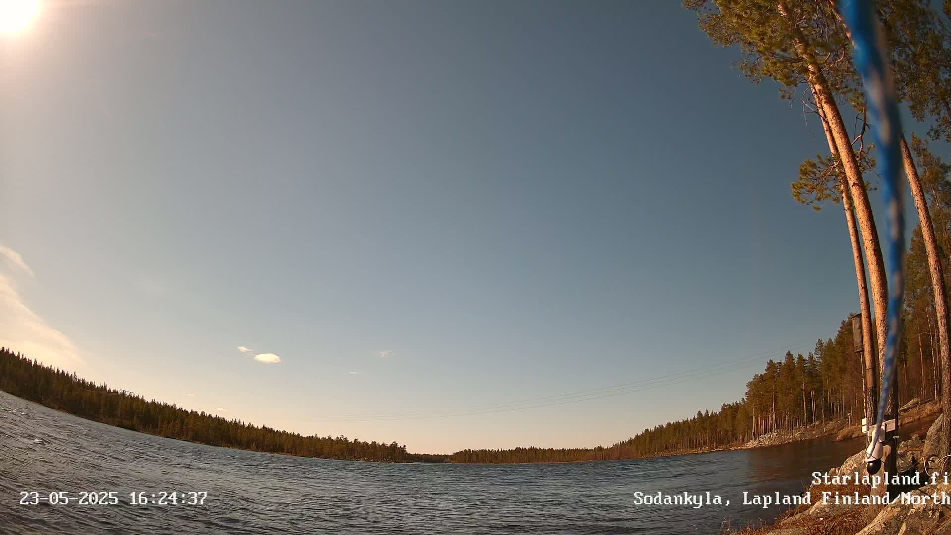 A mostly sunny day shows a calm lake surrounded by a coniferous forest under a clear blue sky.