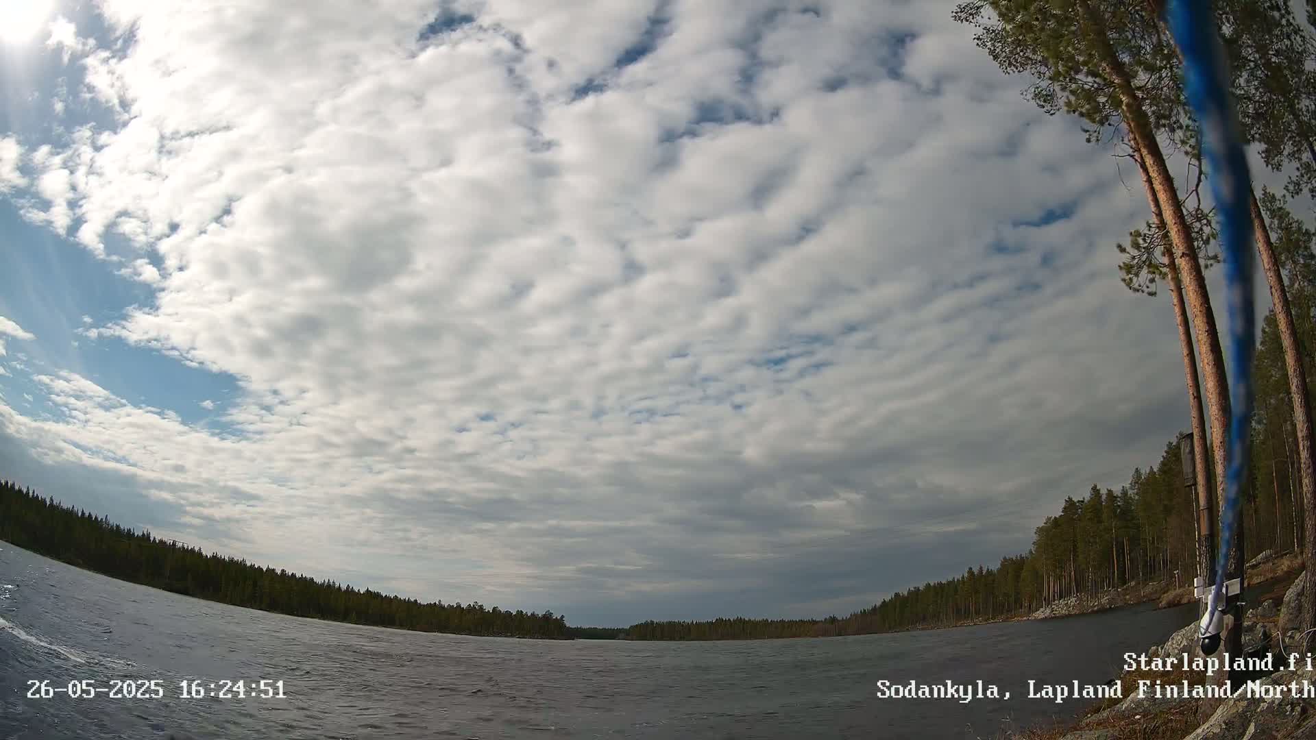 A mostly cloudy sky over a calm lake surrounded by a coniferous forest.