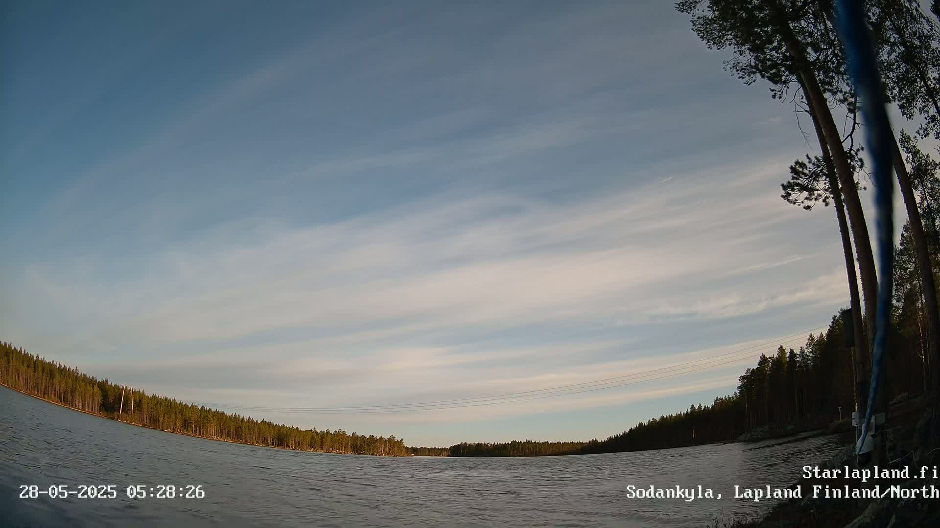 A calm body of water is surrounded by a dark green forest under a mostly clear sky with wispy clouds.