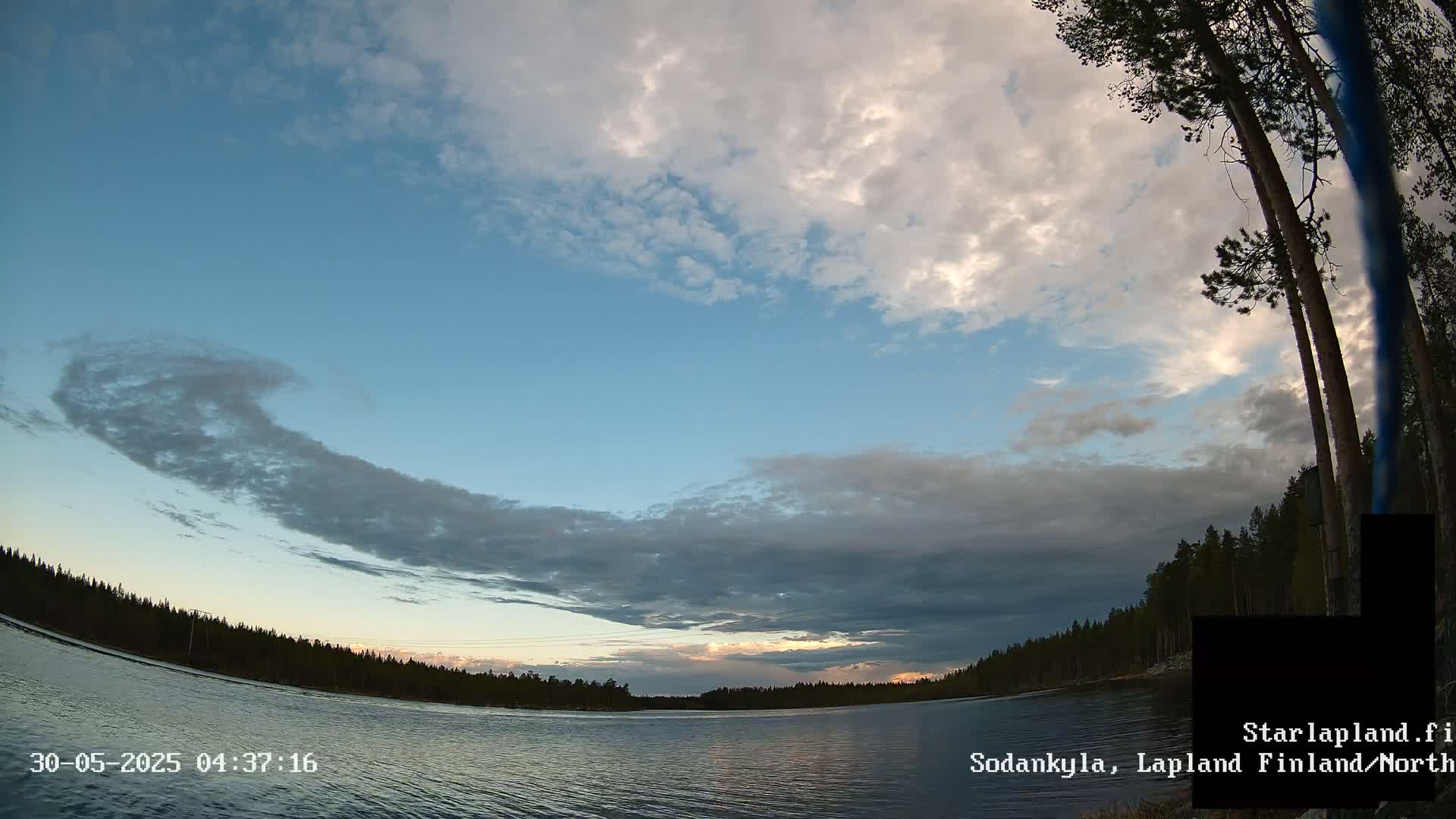 A calm lake is surrounded by a dark green forest under a partly cloudy sky with a mix of blue and gray clouds at sunset.