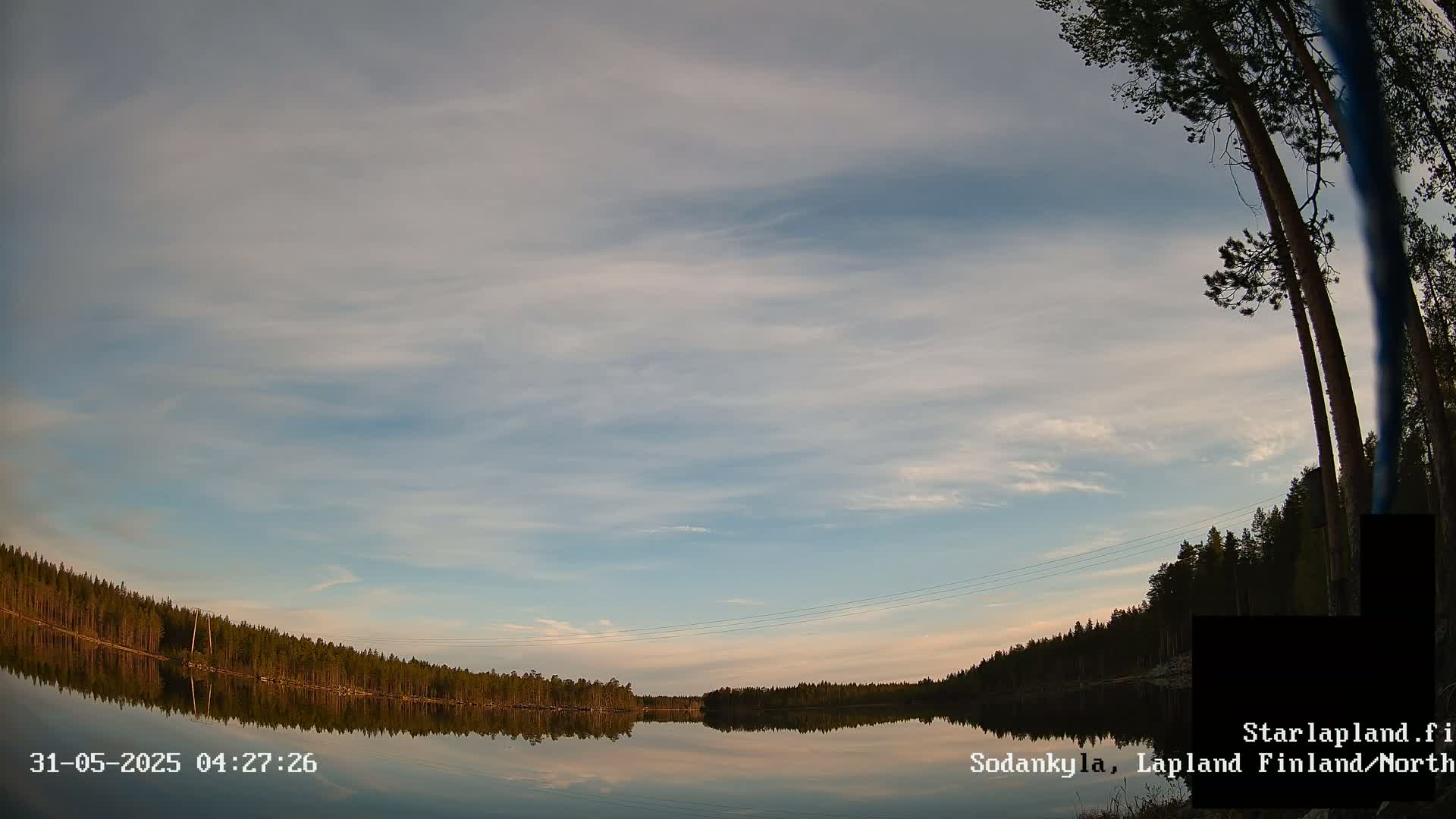 A calm lake reflects a pale sky with thin clouds and a treeline at either edge, under a mostly clear sky.