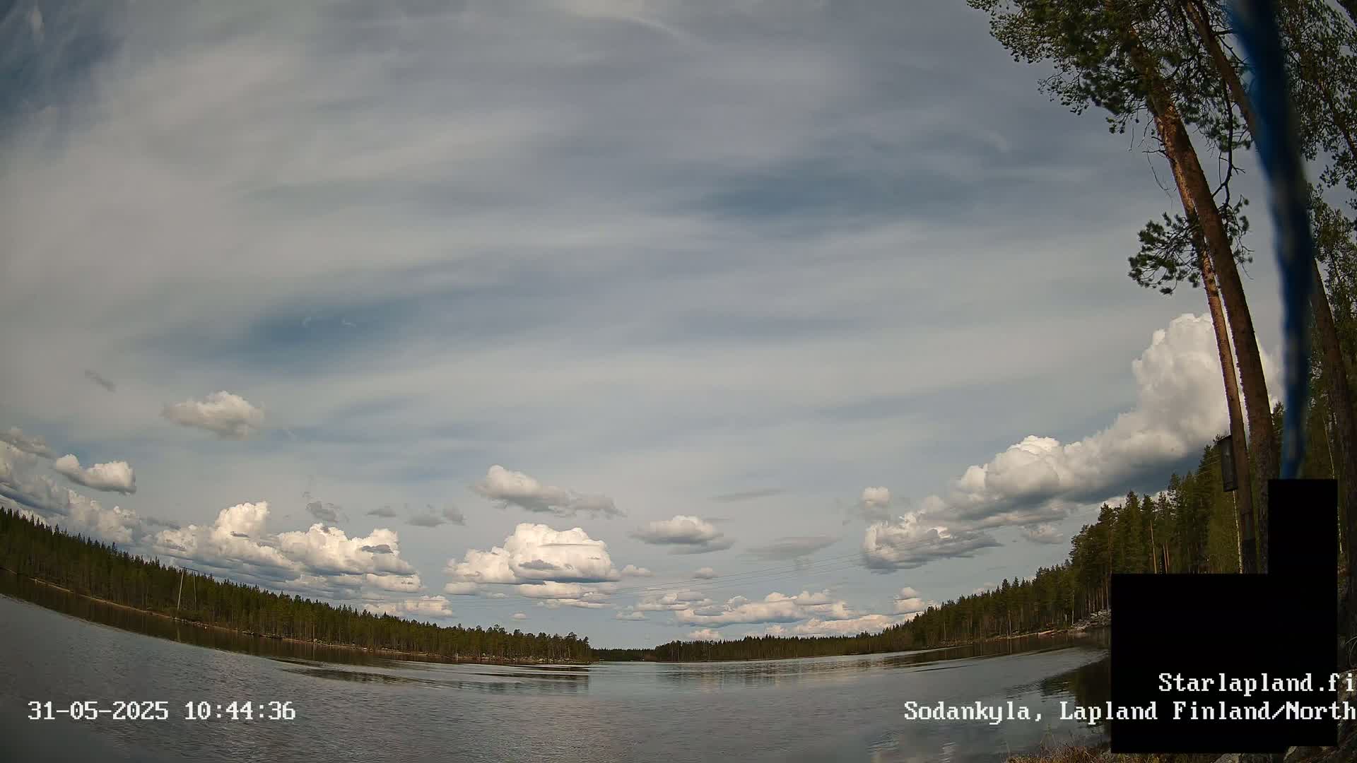 A calm lake is surrounded by a dark green forest under a partly cloudy sky.
