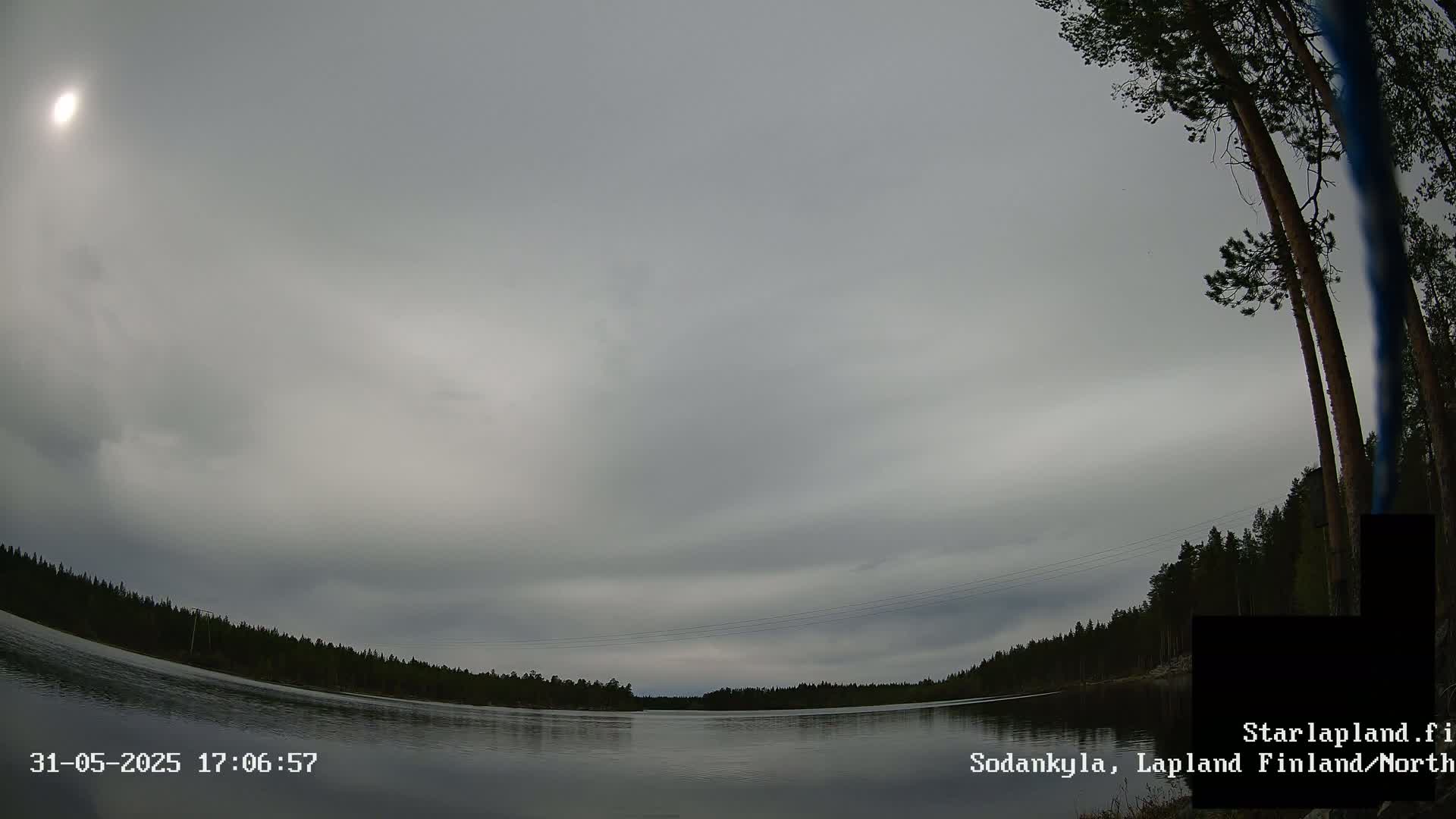A calm, overcast body of water is surrounded by a dark, coniferous forest under a gray sky.