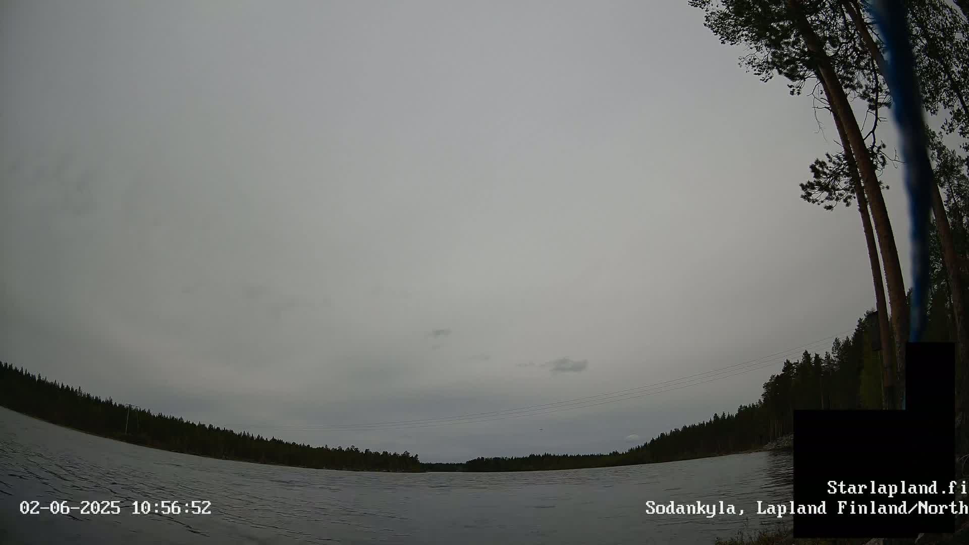 A wide, overcast sky hangs over a calm lake bordered by dark evergreen trees.
