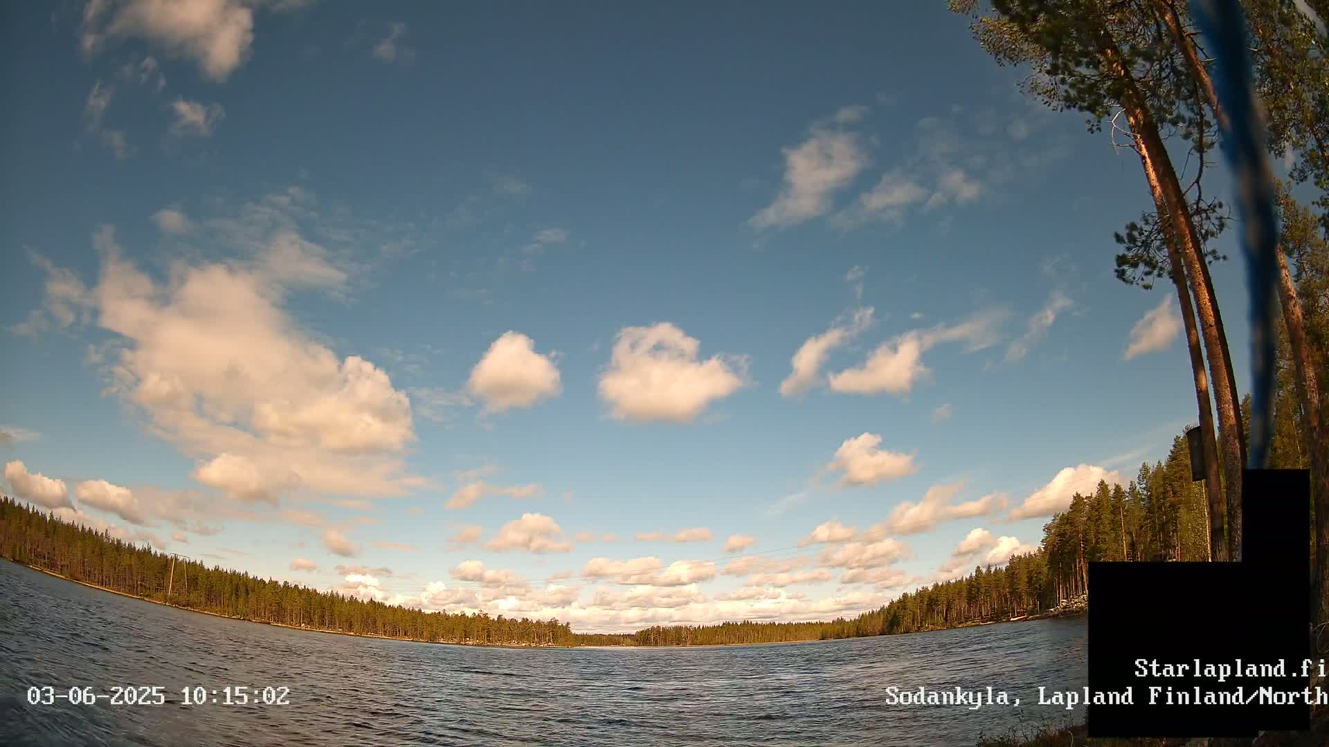 A calm lake is surrounded by a pine forest under a partly cloudy sky.