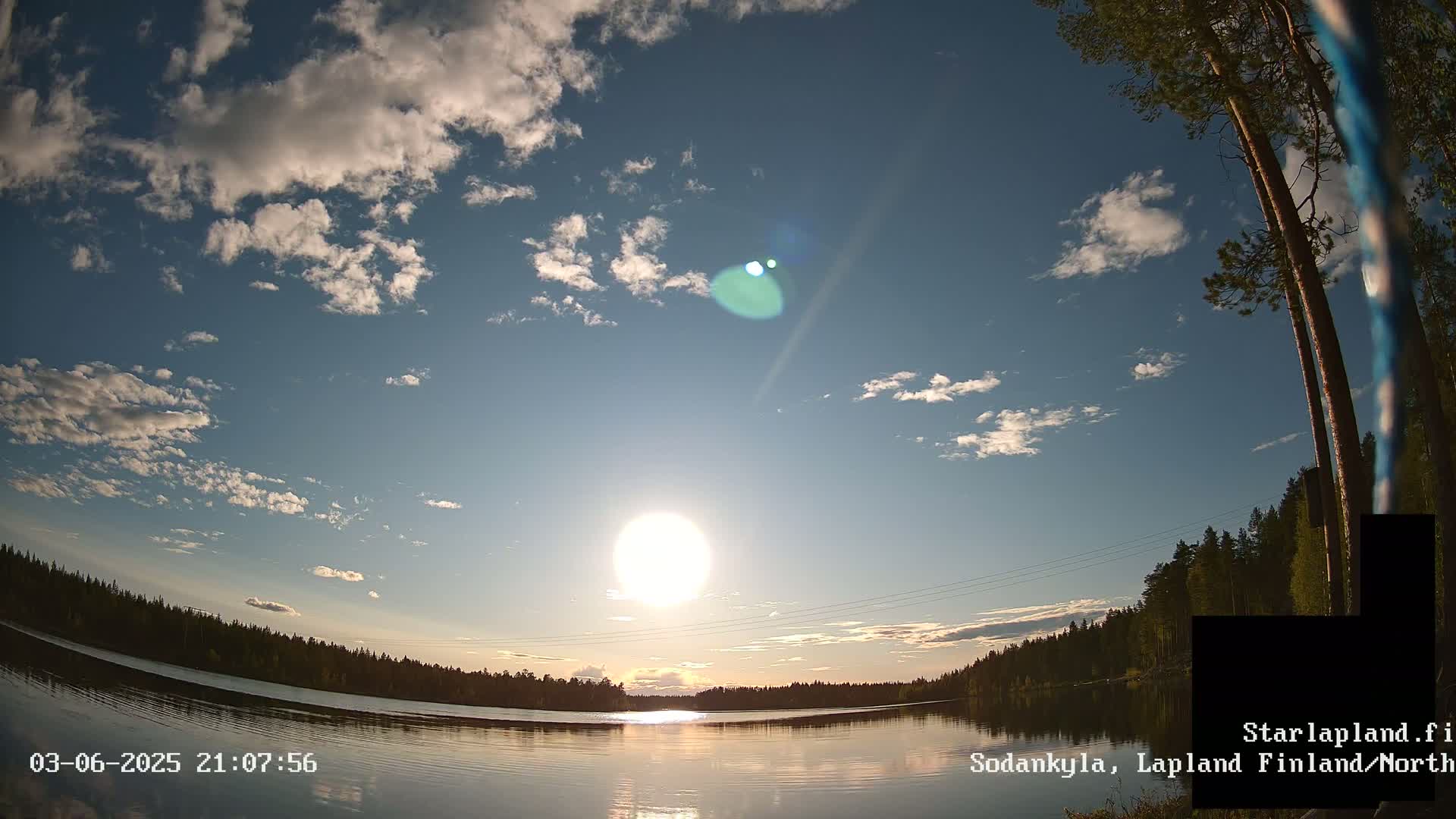 A bright sun shines in a partly cloudy sky over a calm lake bordered by dark green trees.