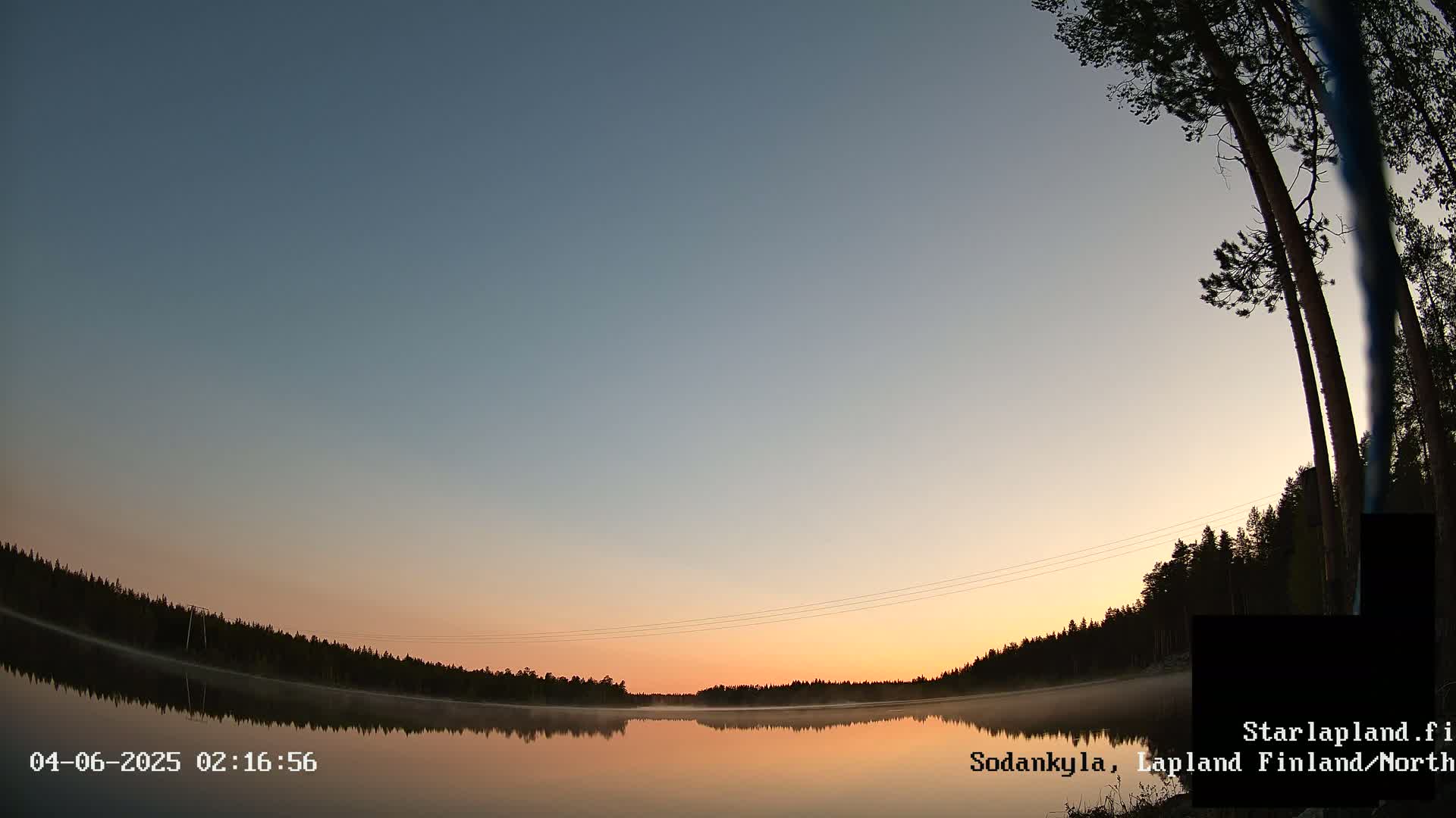 A calm lake reflects a sunrise over a dark forest under a clear, twilight sky.
