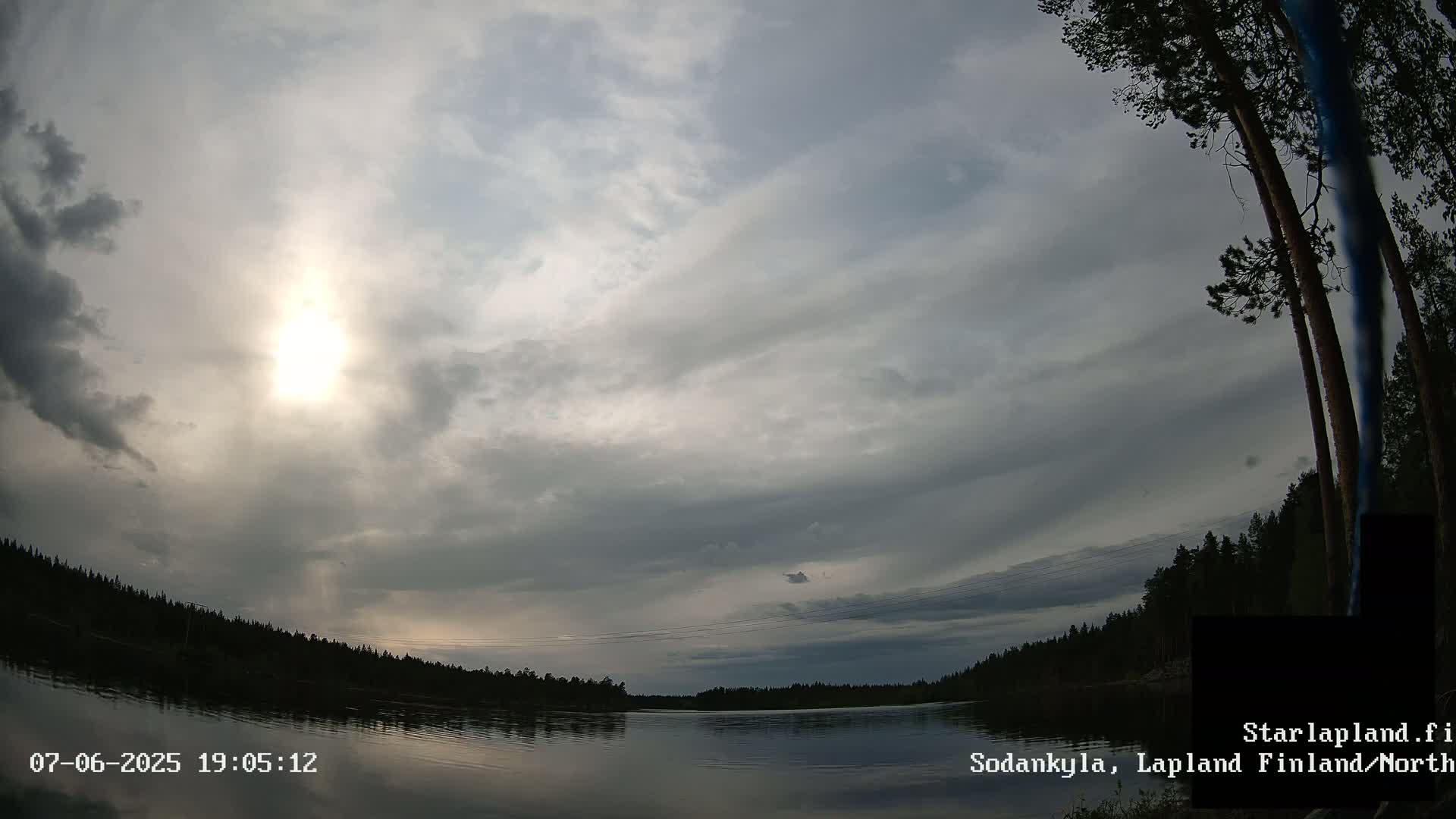 A partly cloudy sky with the sun shining through, over a calm lake surrounded by a dark line of trees.