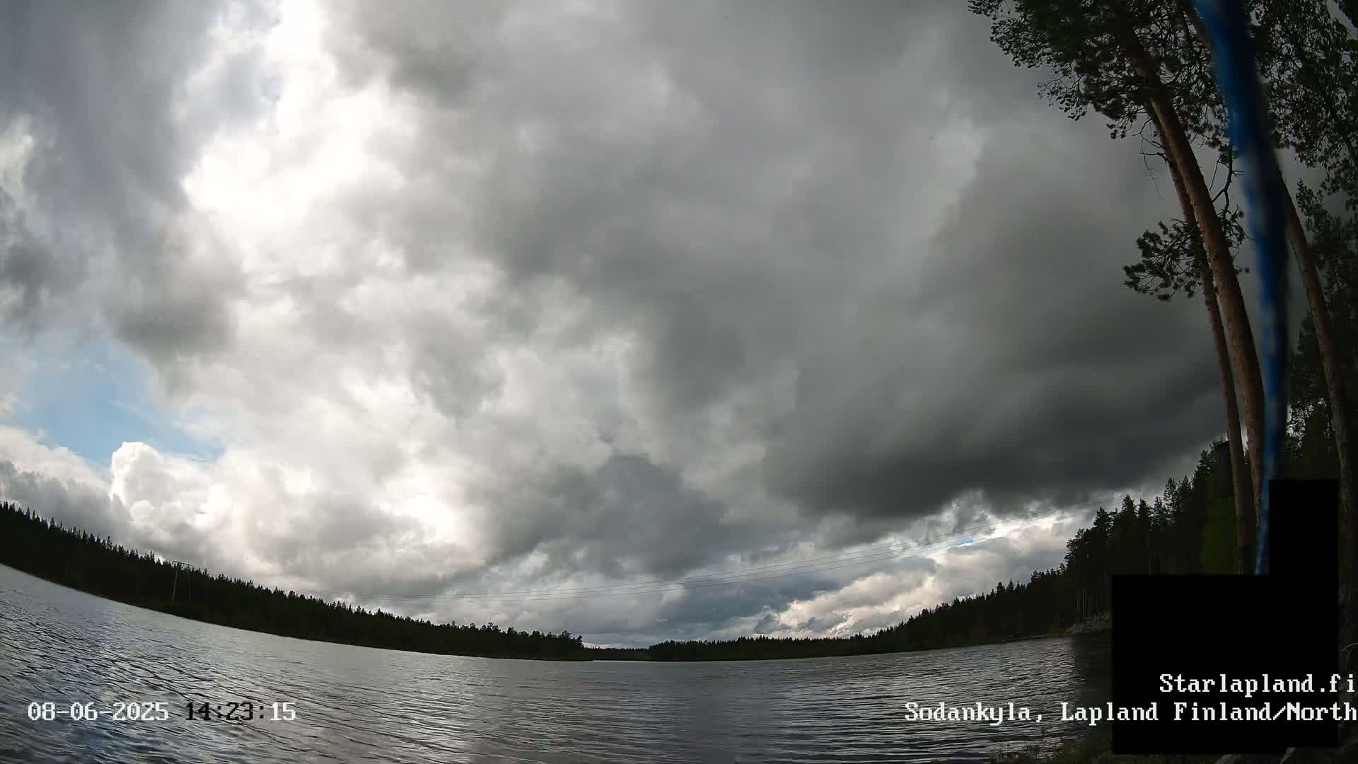 A mostly cloudy sky hangs over a calm lake surrounded by dark green trees.