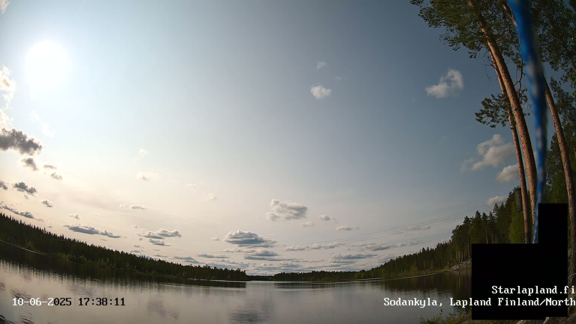 A calm lake reflects a mostly sunny sky with scattered clouds, surrounded by a dark green forest under a partly sunny sky.