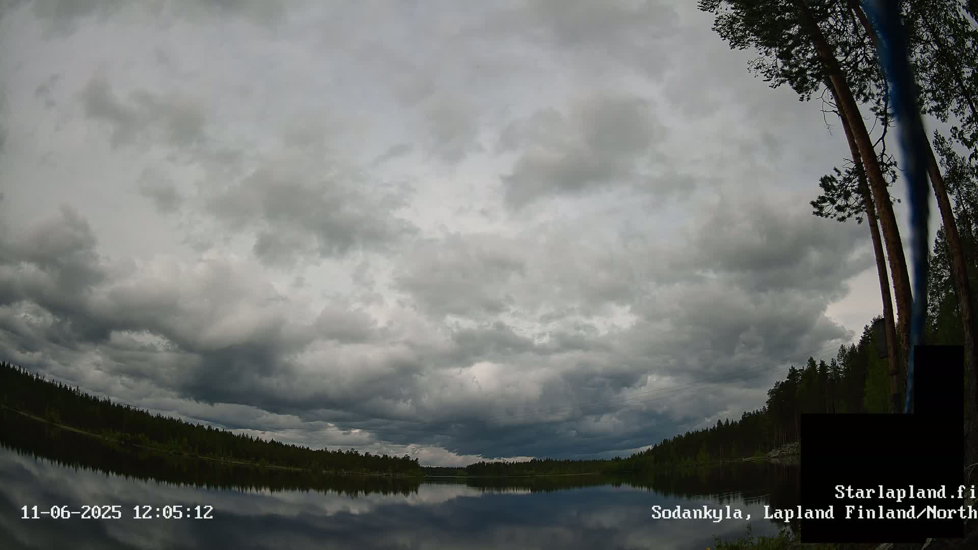 A calm lake reflects a heavily overcast sky under which dark, coniferous trees line the shoreline.