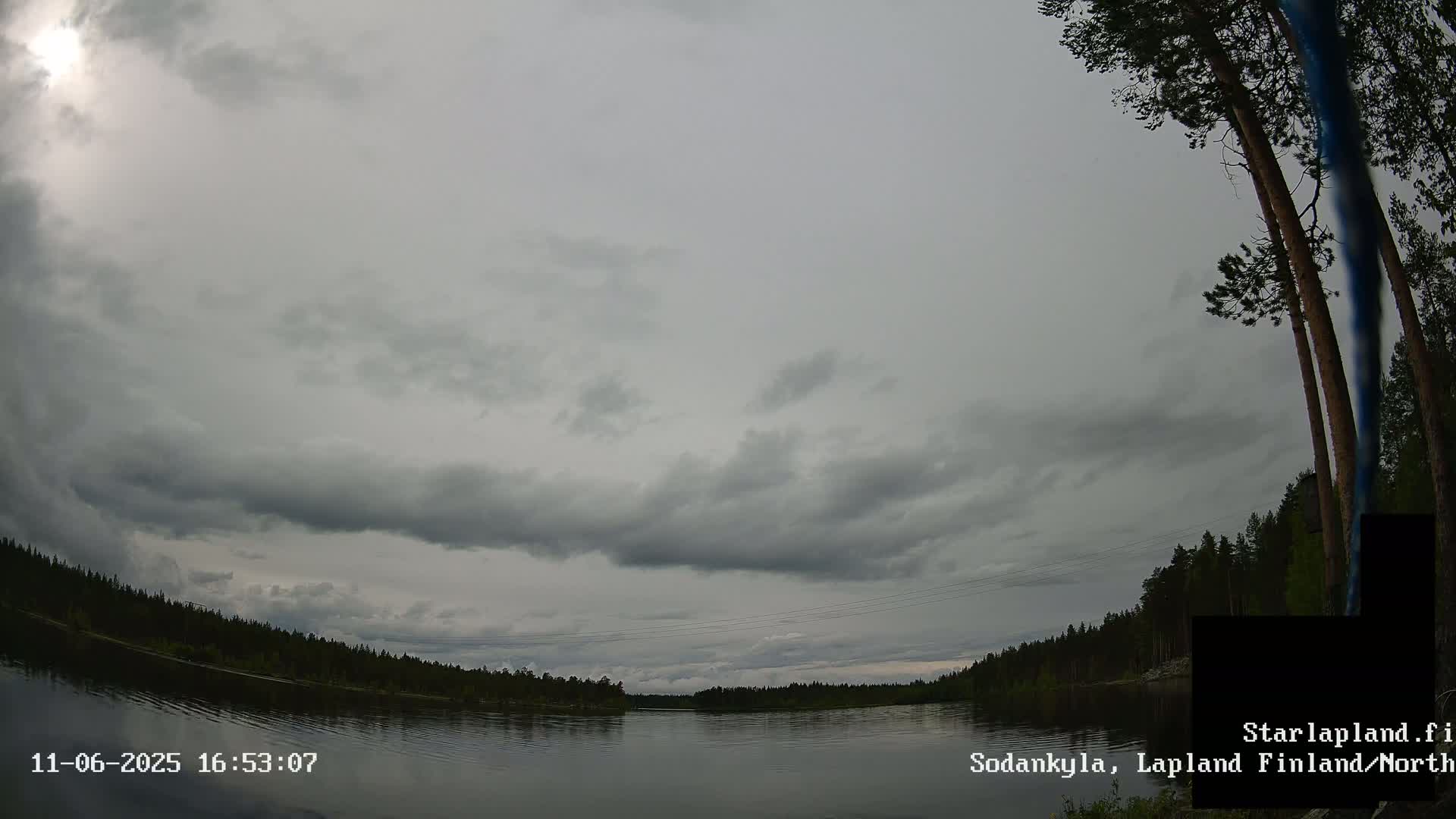 A mostly cloudy sky hangs over a calm lake bordered by dark green trees.