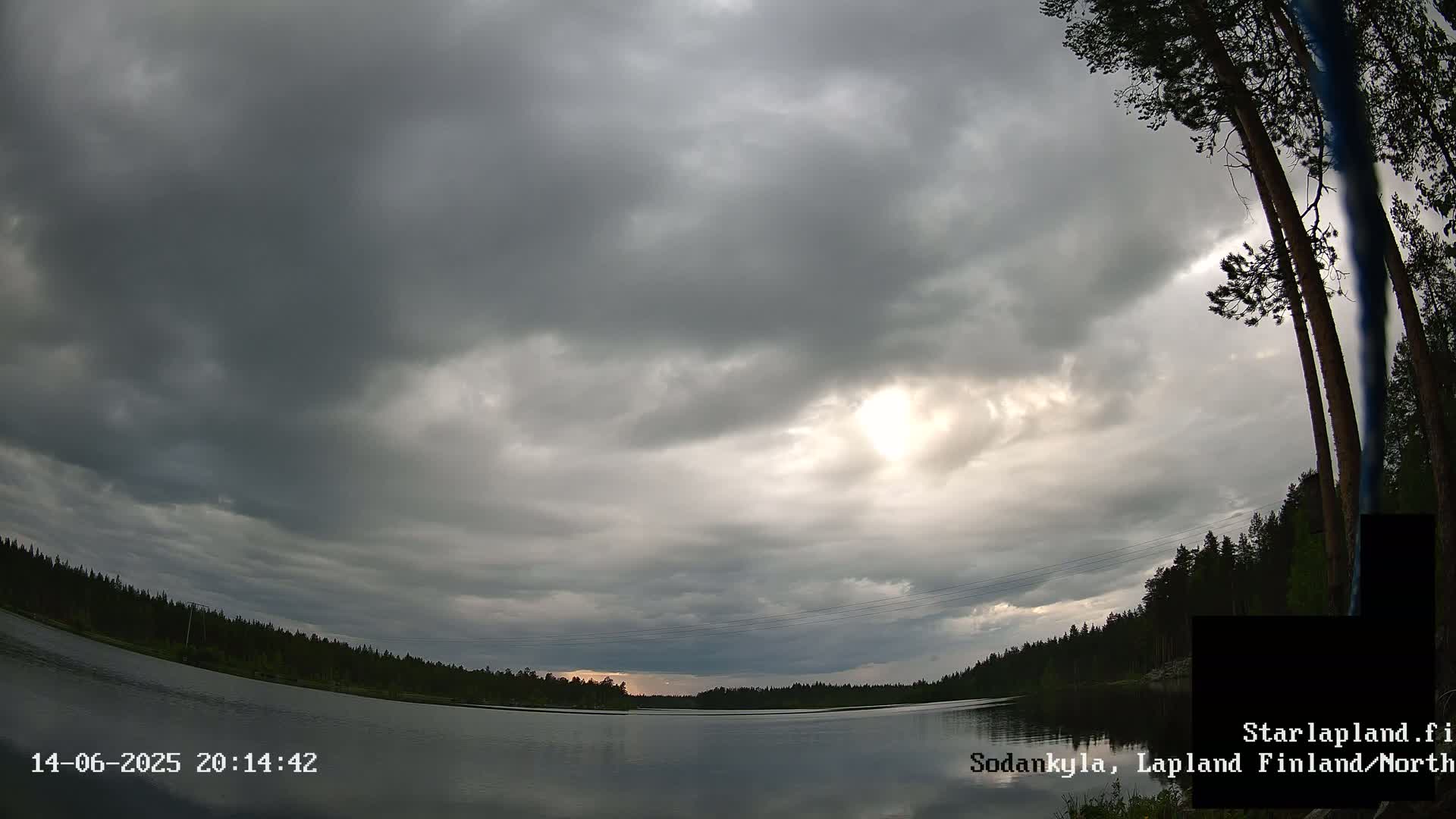 A calm lake is surrounded by a dark, overcast sky and a silhouetted forest under a mostly cloudy sky.