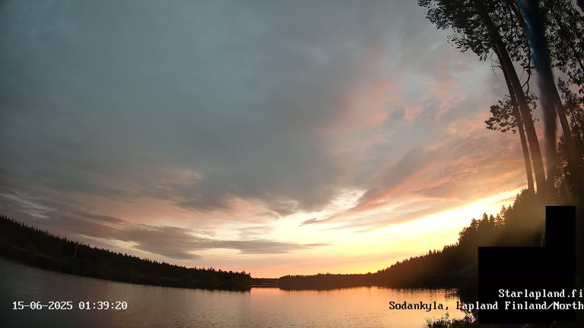 A calm lake is surrounded by a dark, overcast sky and a silhouetted forest under a mostly cloudy sky.
