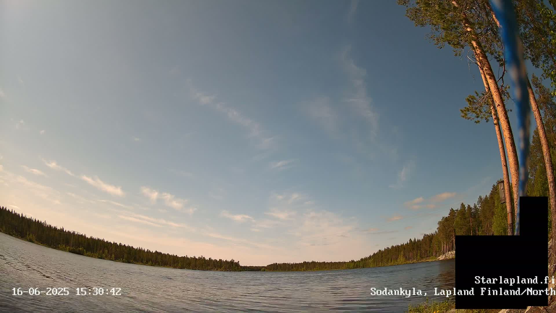 A calm lake is surrounded by a coniferous forest under a mostly clear, light blue sky with wispy clouds.