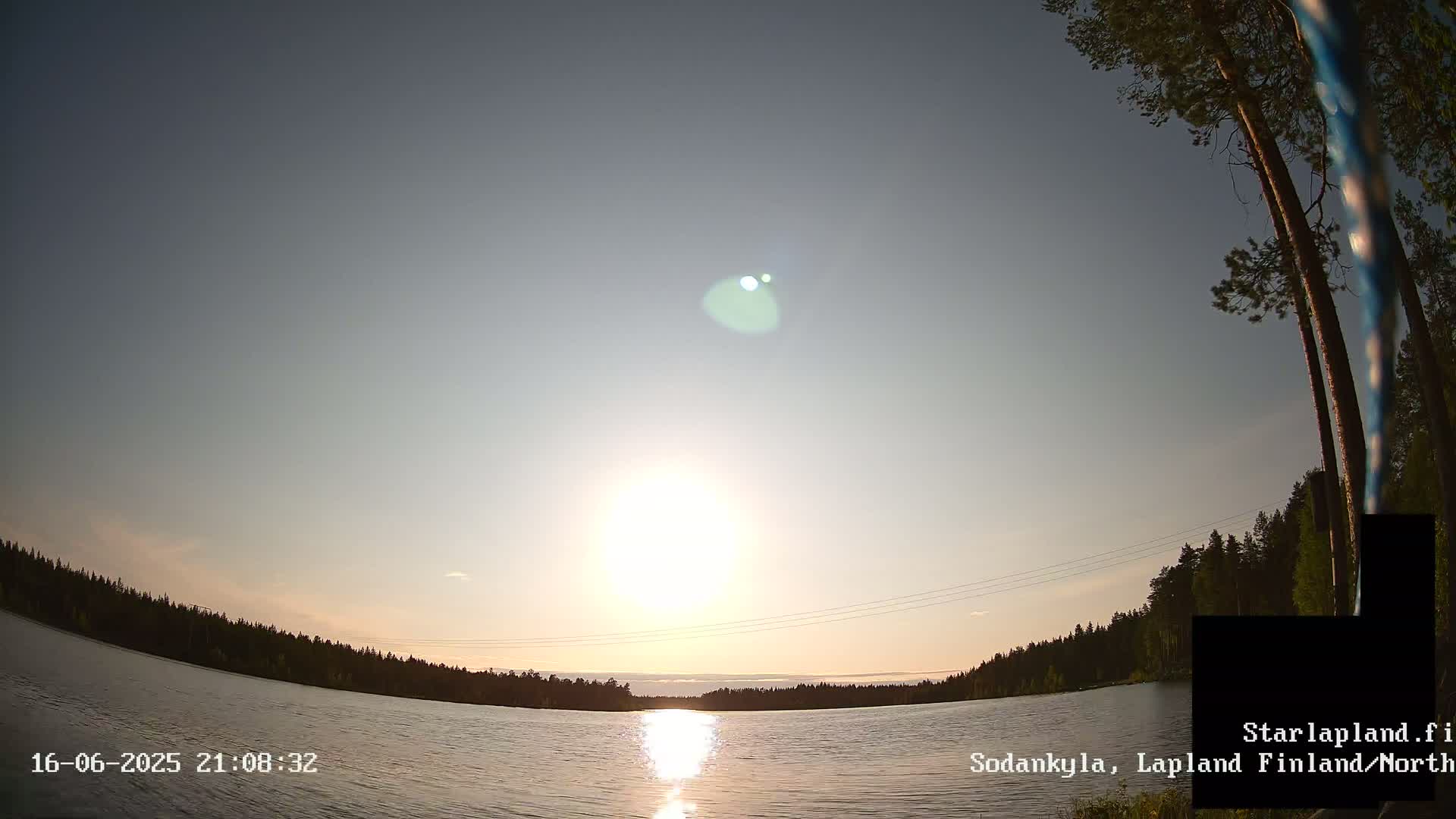 A calm lake reflects a bright sun setting over a dark treeline under a clear sky.
