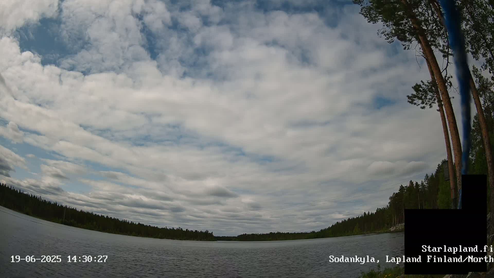 A mostly cloudy sky overlooks a calm lake surrounded by a dark green forest.