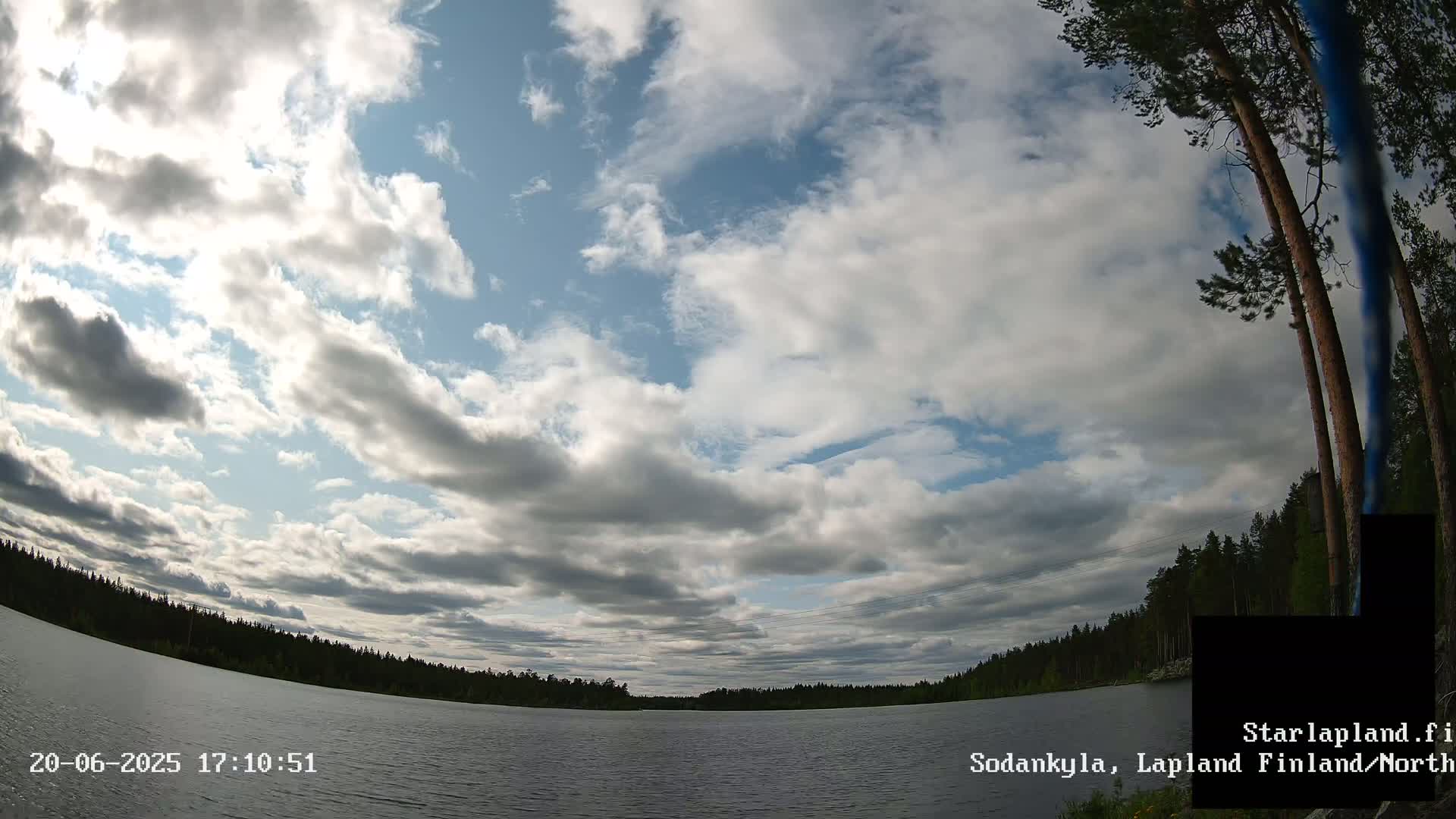 A partly cloudy sky overlooks a calm lake surrounded by a dark green forest.