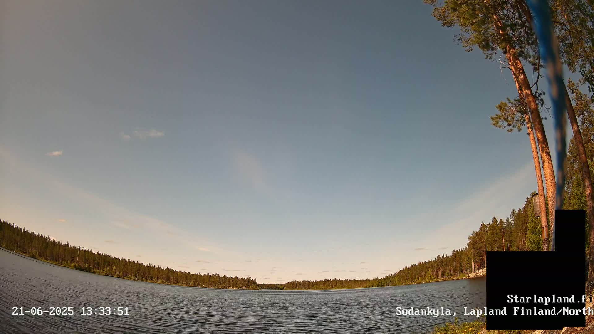 A calm lake is surrounded by a pine forest under a mostly clear, light-blue sky.