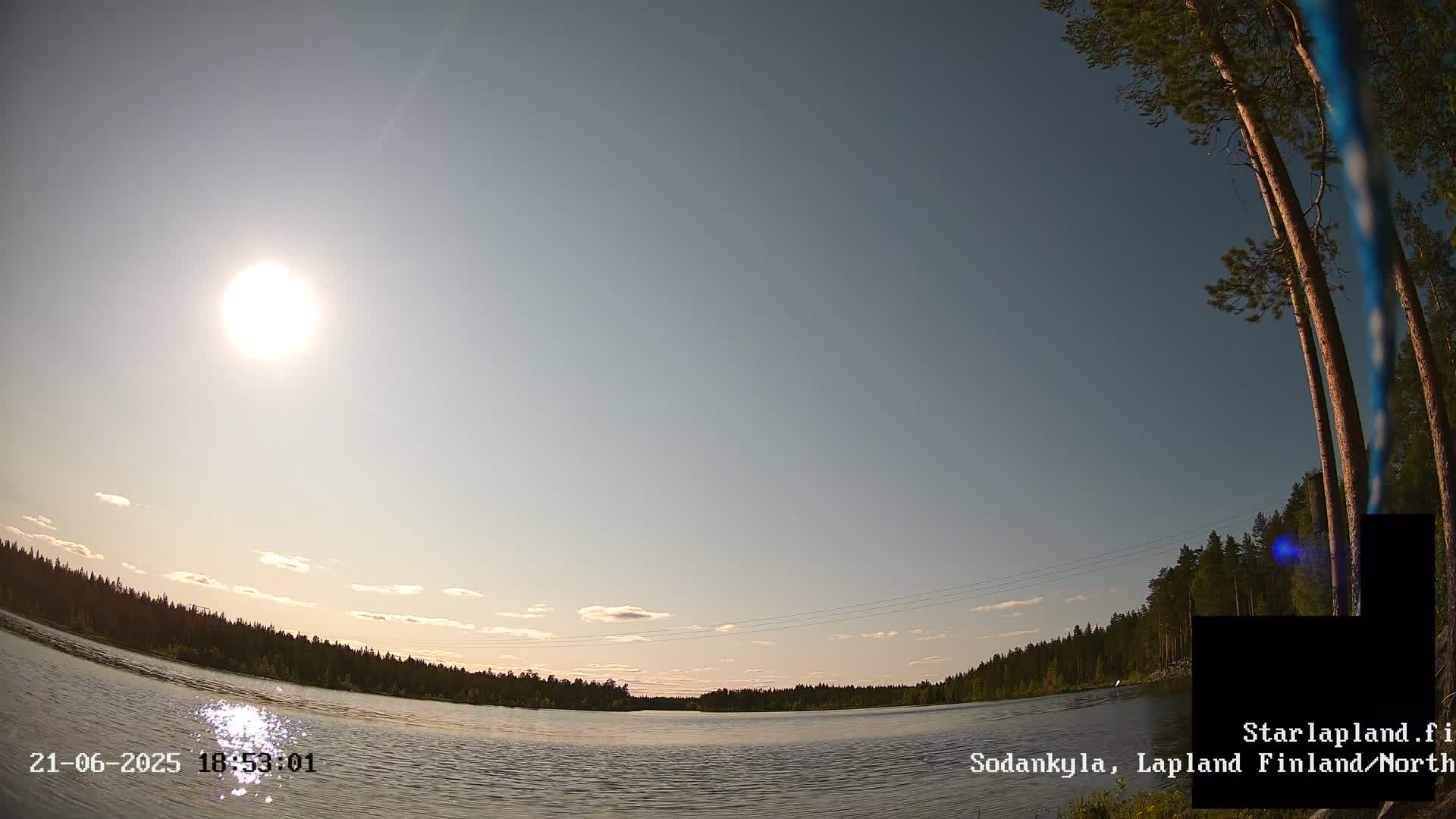 A sunny, mostly clear sky overlooks a calm lake bordered by a dark green forest.