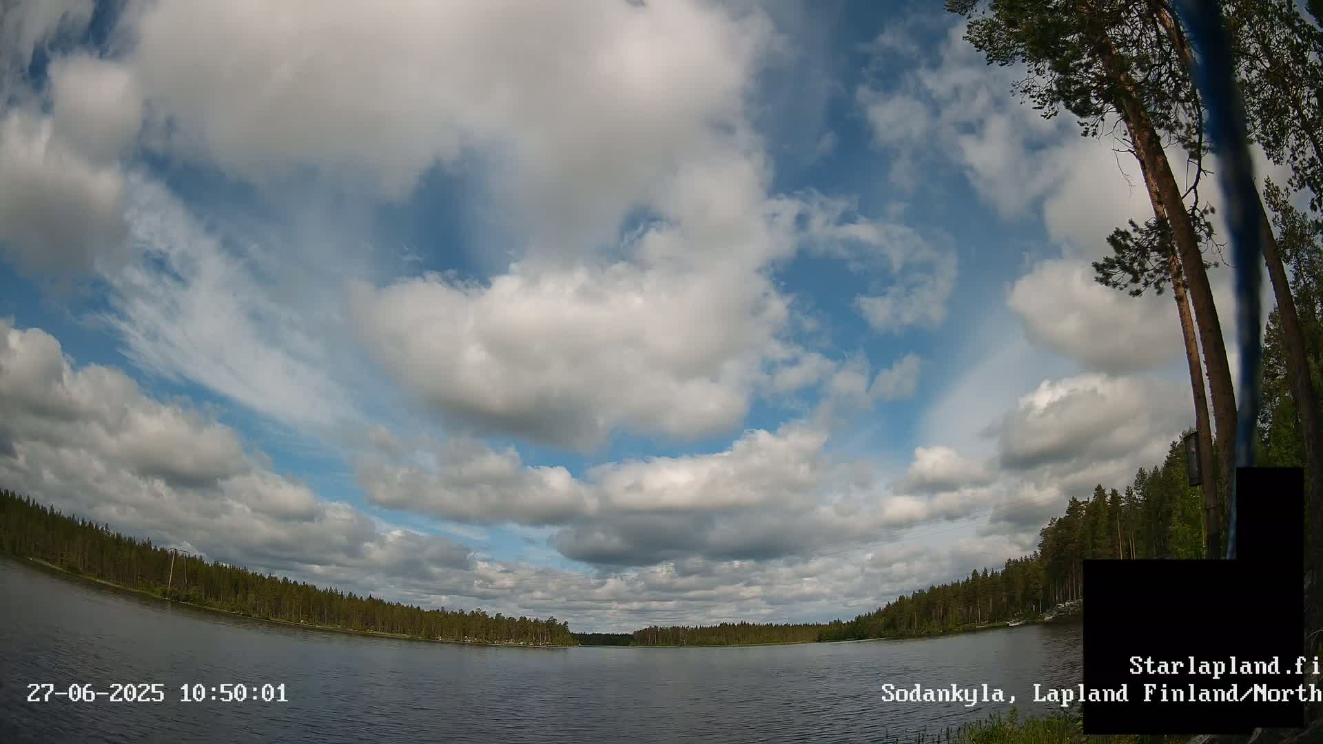 A calm lake is surrounded by a pine forest under a partly cloudy sky.