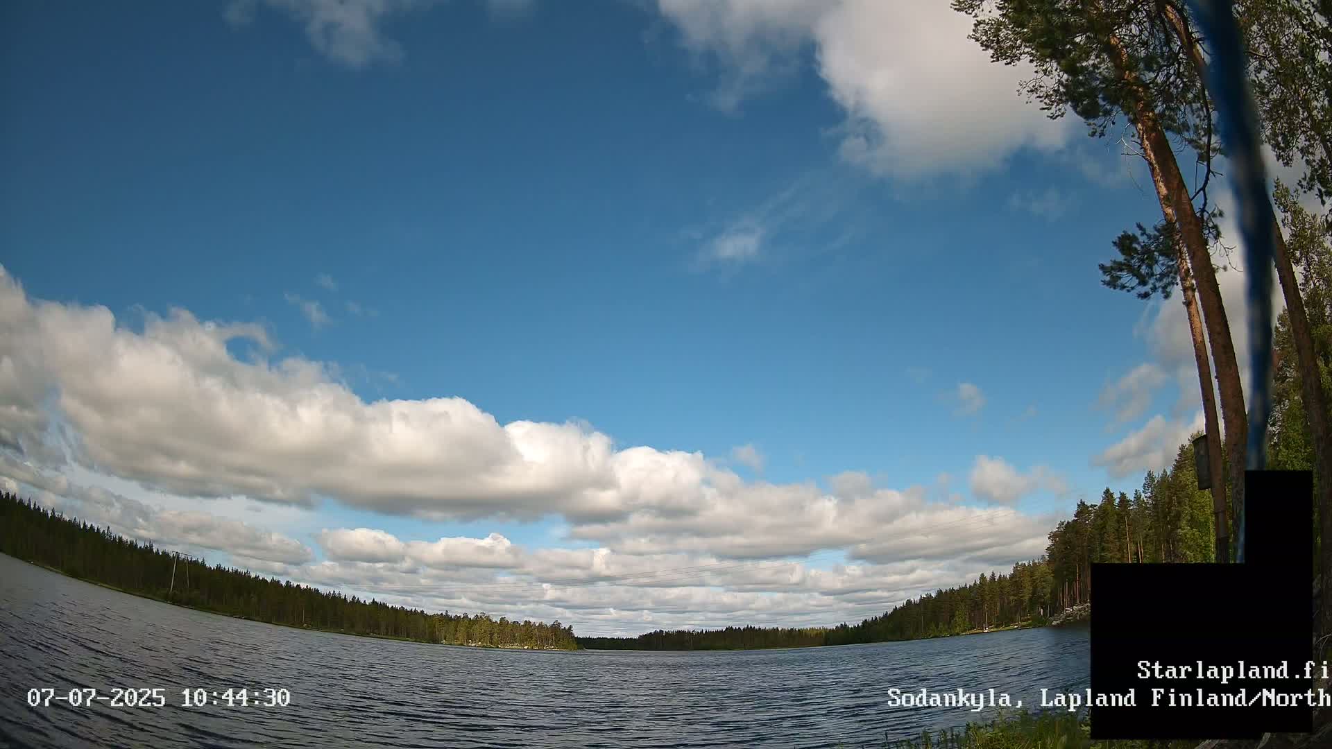 A calm lake is surrounded by a dense forest under a mostly sunny sky with fluffy white clouds.