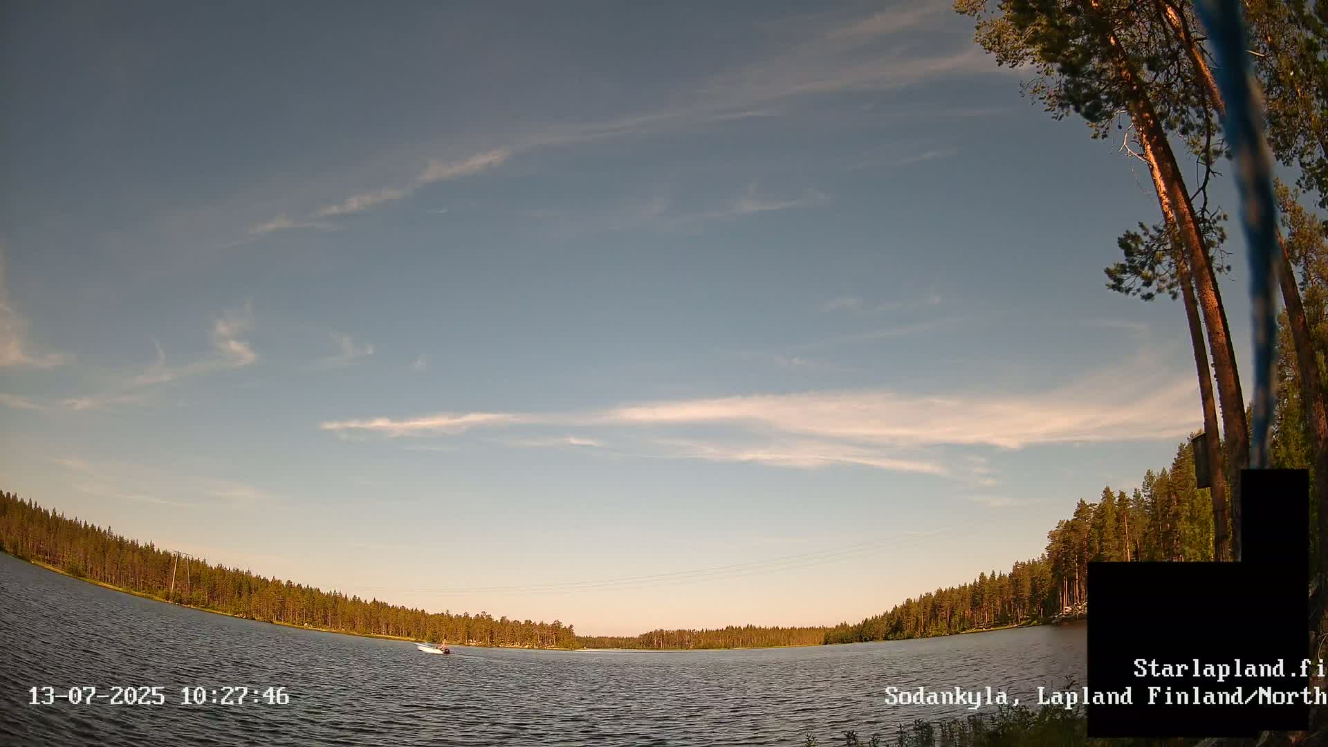 A calm lake surrounded by a coniferous forest under a mostly clear, light-blue sky with a few wispy clouds.