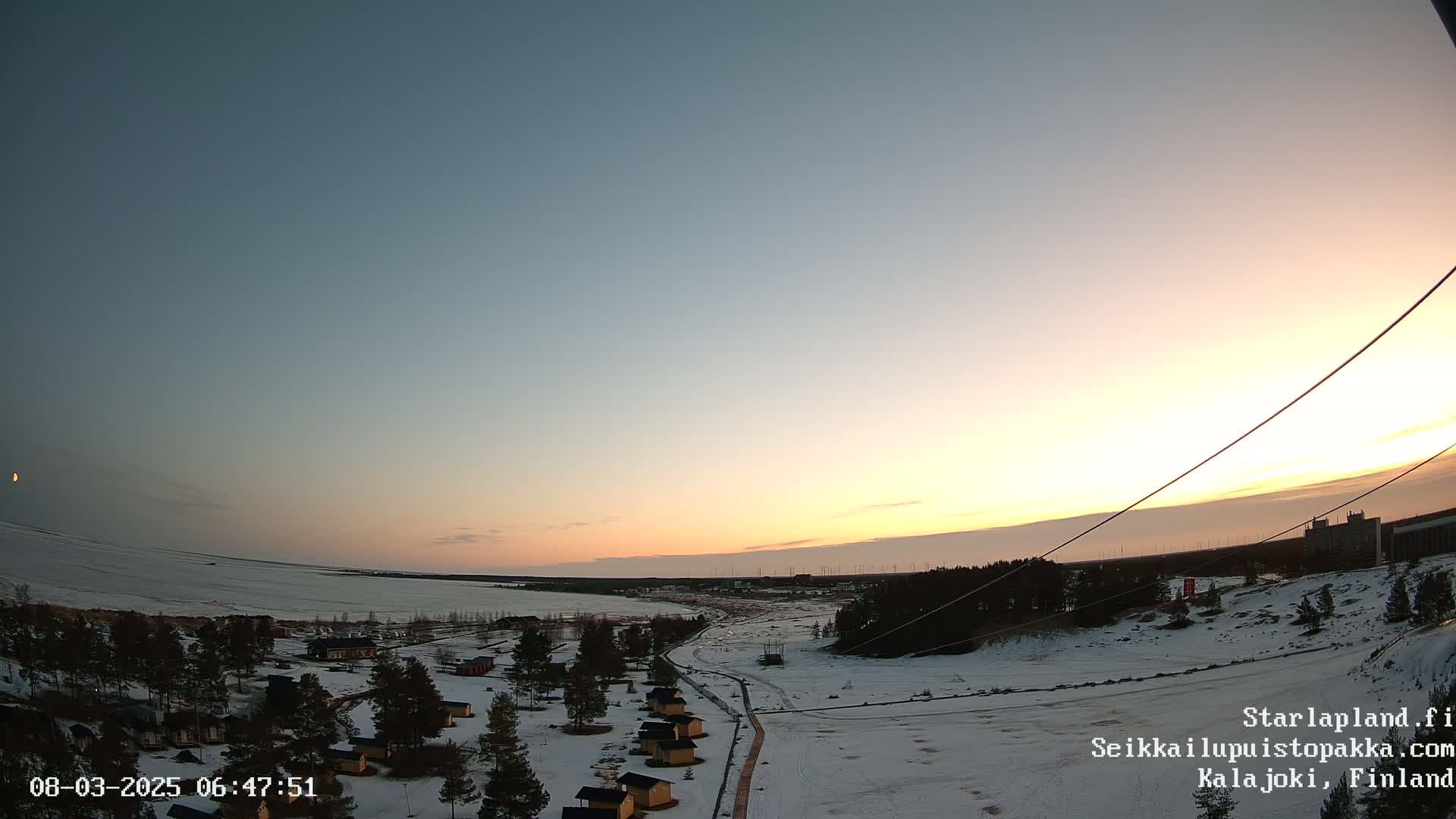 A snow-covered landscape with small cabins at sunrise under a clear, pale-orange and blue sky.