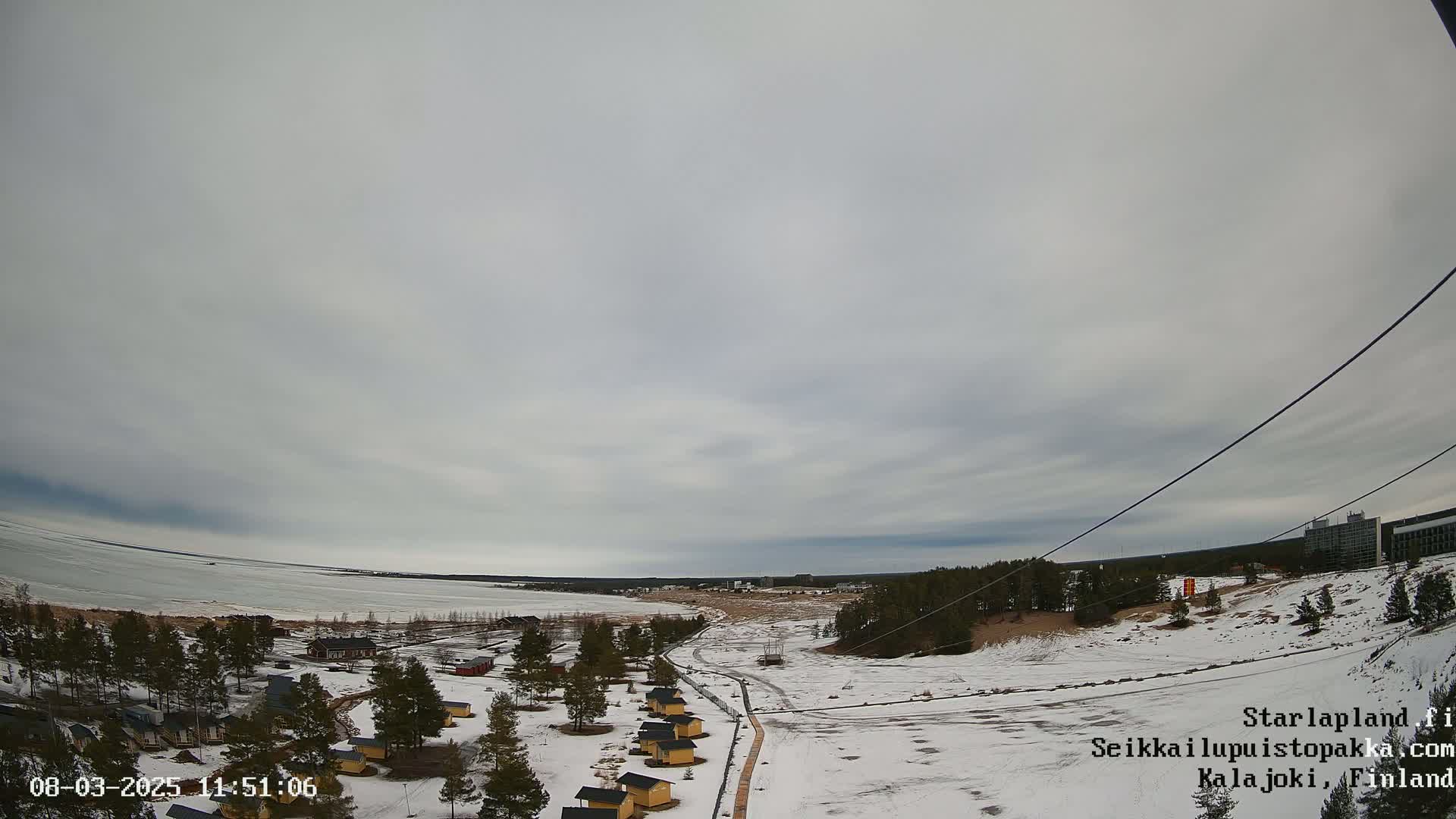 A mostly cloudy day reveals a snow-covered landscape with a frozen lake, small cabins, and a distant building, viewed from a high vantage point.